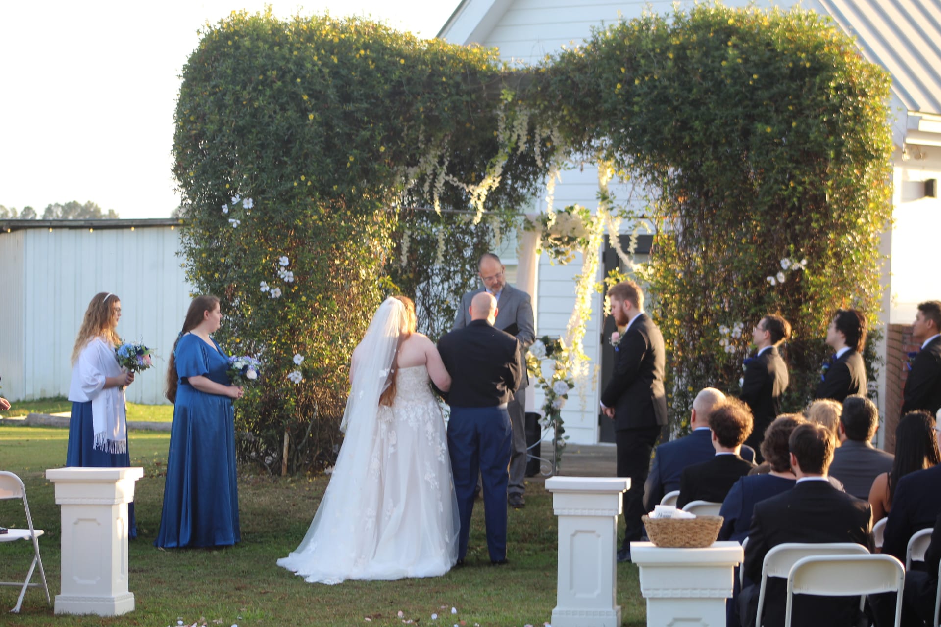 Bride and groom exchange vows beneath a lush green floral arch on the ceremony lawn at Scarborough House, surrounded by bridesmaids, groomsmen, and seated guests.