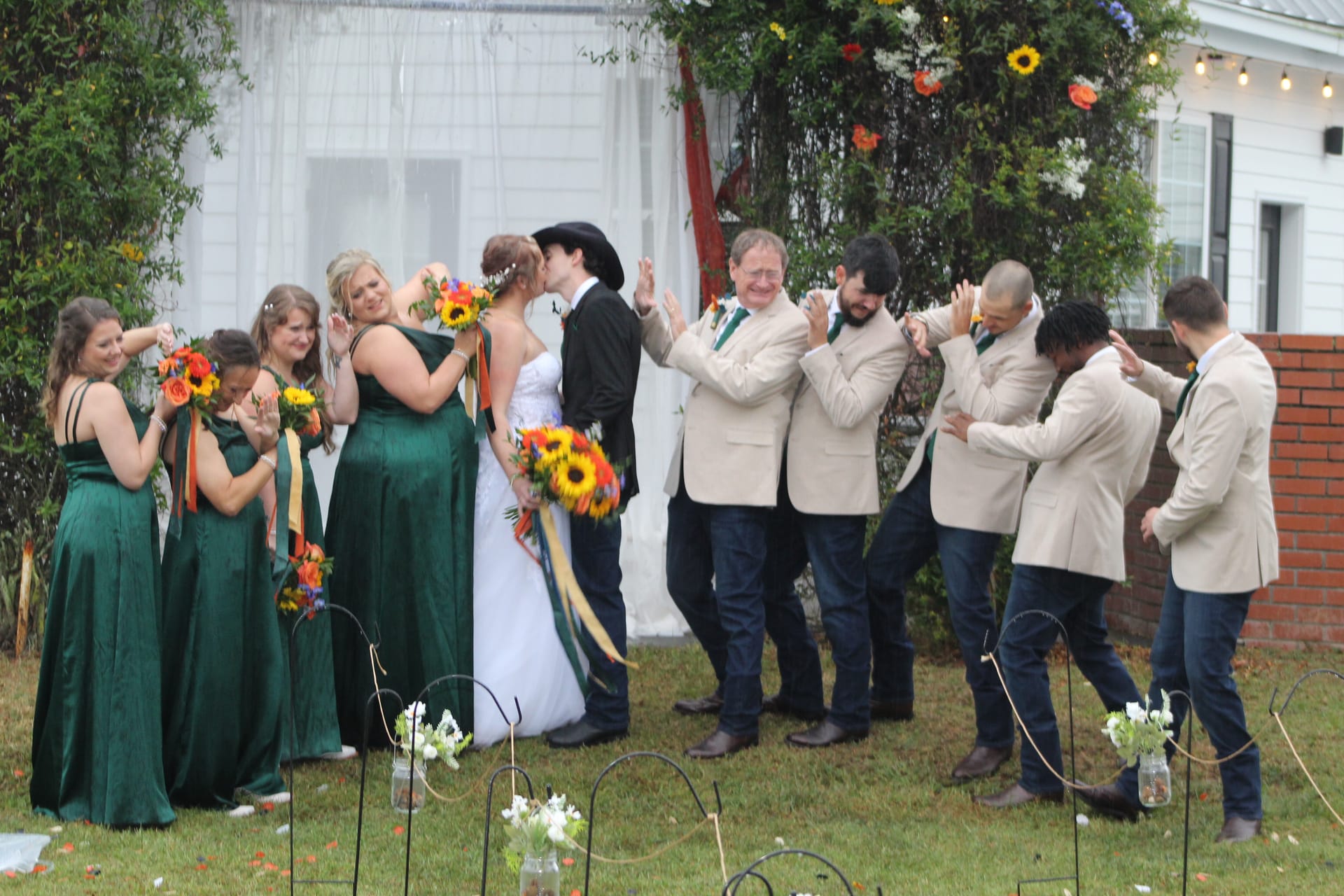 Bride and groom share their first kiss as bridesmaids in emerald green gowns and groomsmen in tan blazers cheer on the ceremony lawn at Scarborough House.