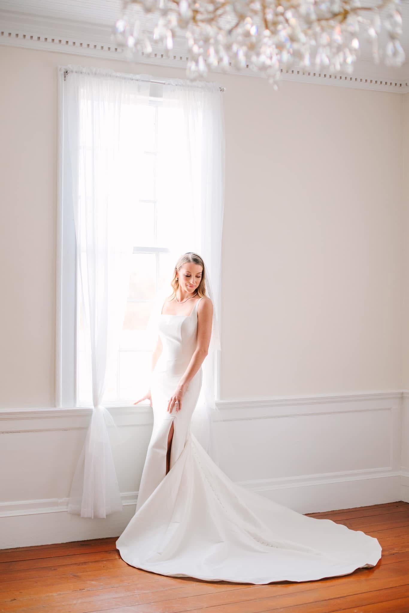 Bride in a sleek white mermaid wedding gown with a long train stands gracefully by a tall sunlit window inside the historic Scarborough House manor, with a crystal chandelier overhead and warm hardwood floors below.