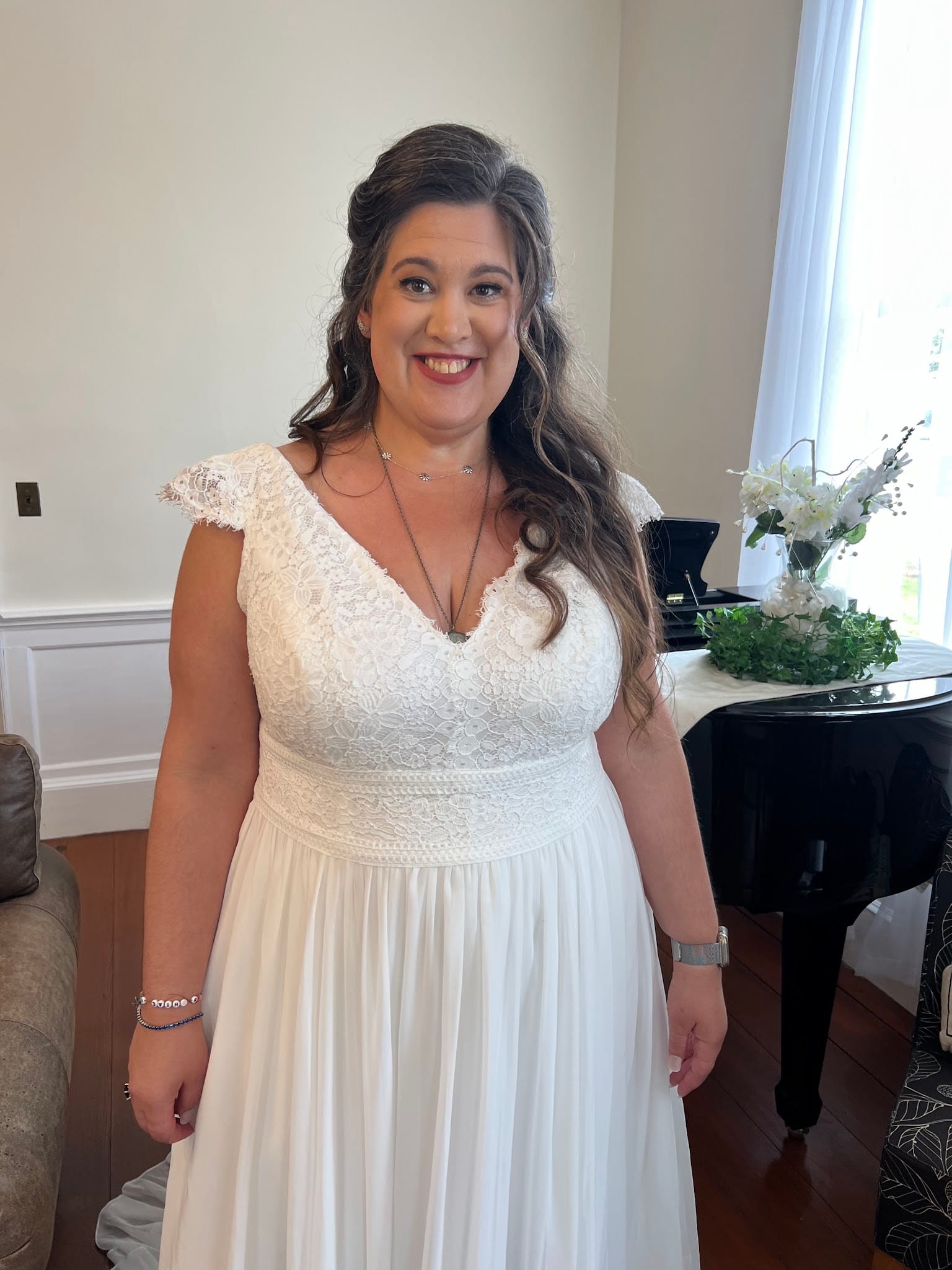 Smiling bride in a white lace cap-sleeve wedding gown poses in an elegant indoor room with a grand piano and white floral arrangement at Scarborough House.
