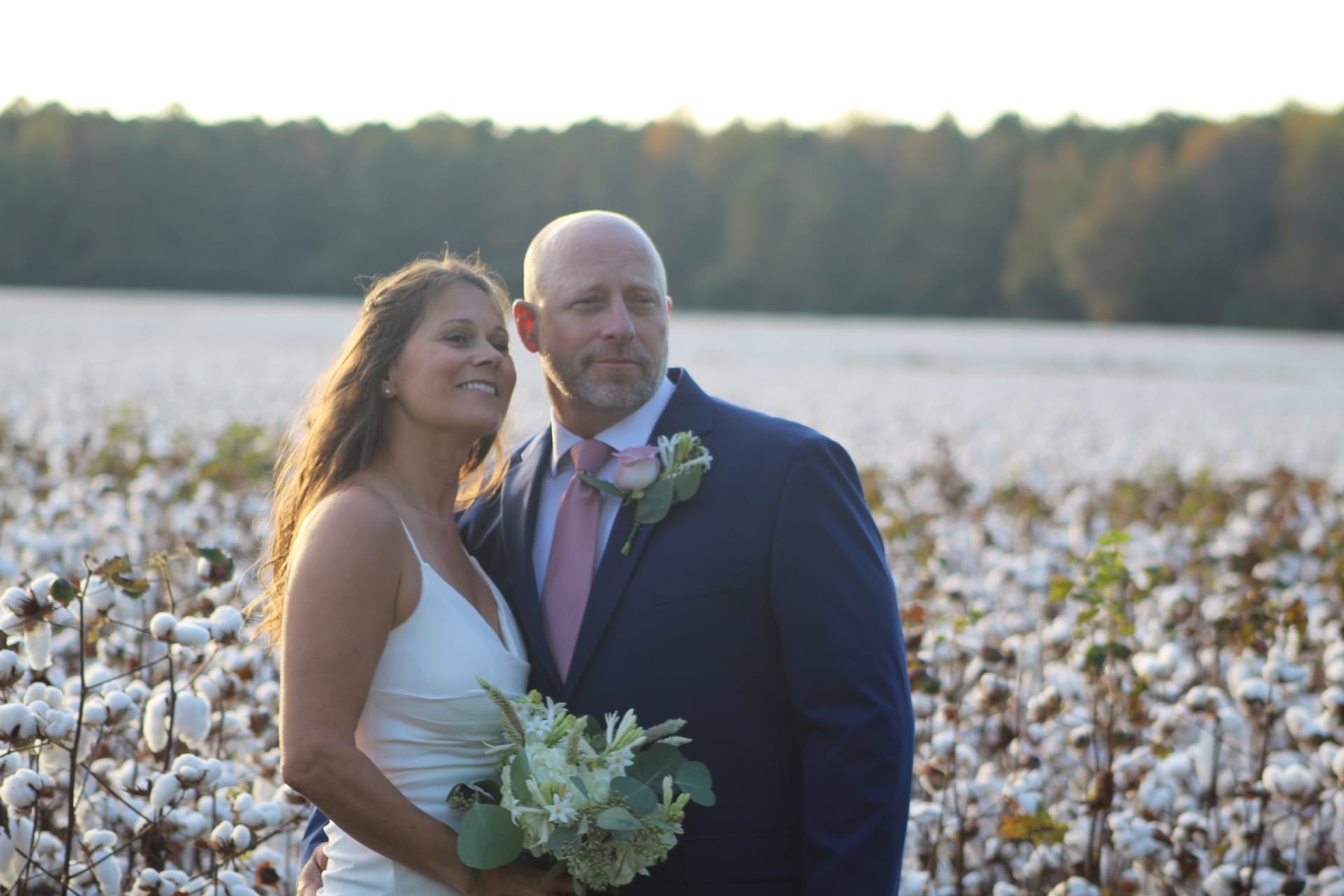 Bride in white gown holding a green bouquet and groom in navy suit with pink tie and rose boutonniere pose together at golden hour in a blooming cotton field at Scarborough House.