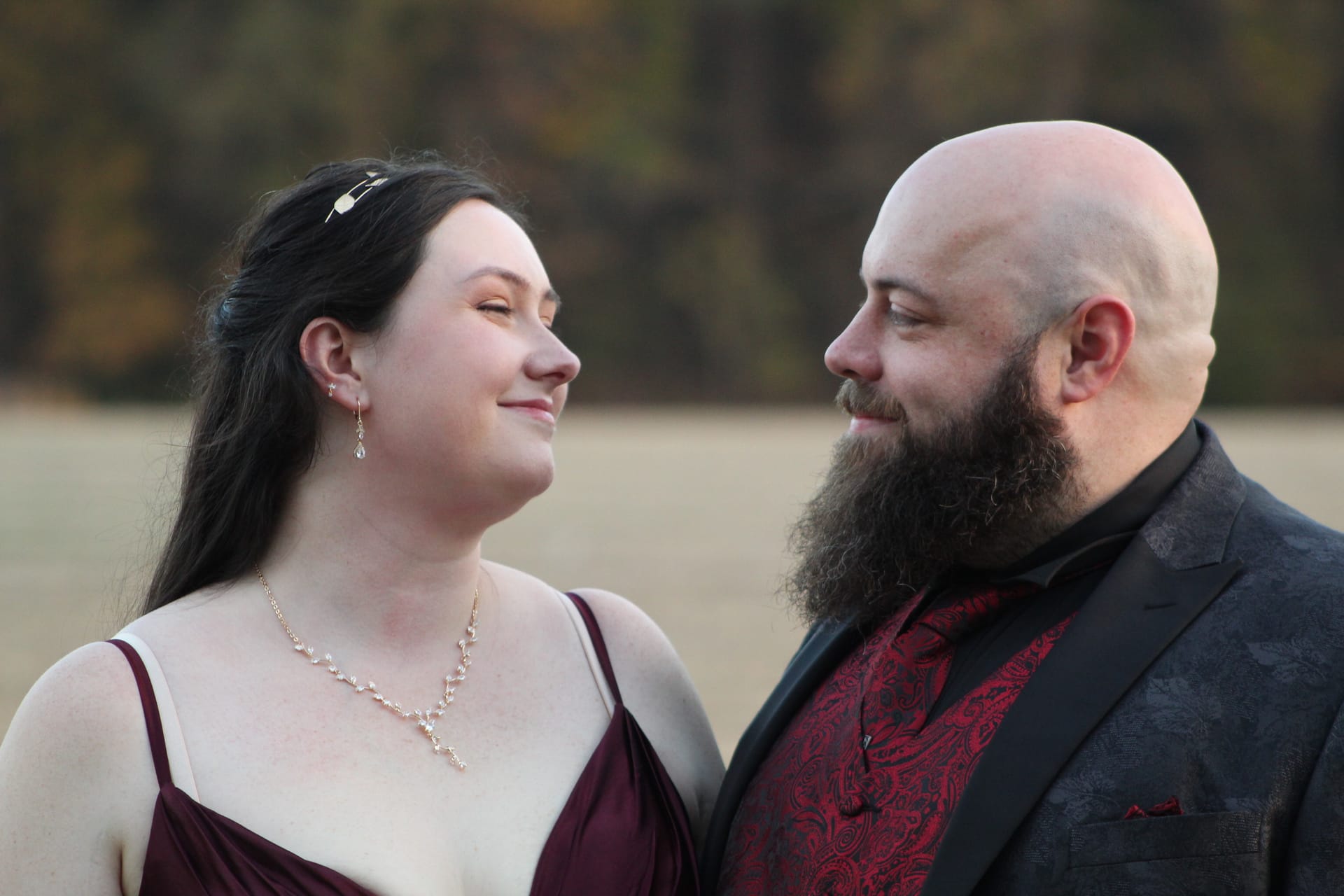 Bride in a burgundy gown and groom in a dark paisley suit with red vest share a tender glance outdoors against a softly blurred autumn farmland backdrop at Scarborough House.