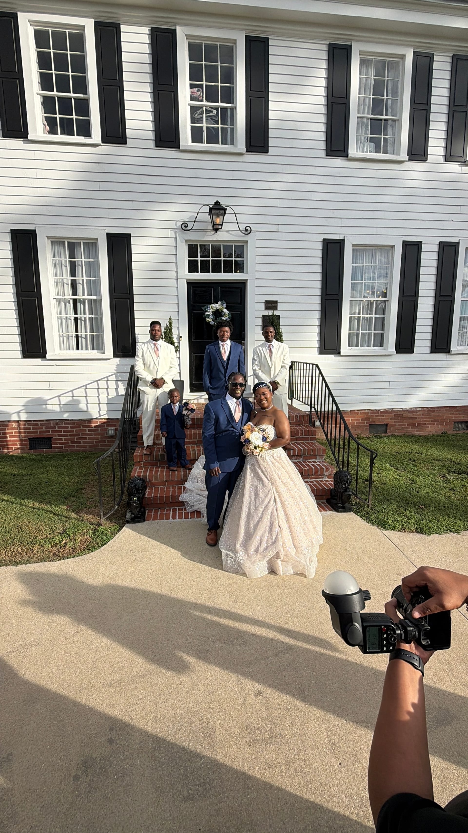 Bride in a strapless lace ballgown and groom in a navy suit pose with their wedding party on the brick front steps of the 1821 Scarborough House manor.