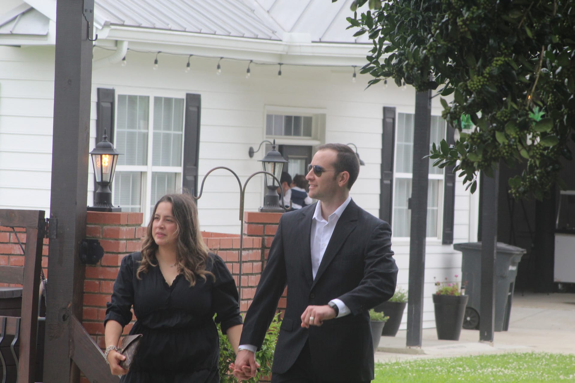 Elegantly dressed couple in black attire walking hand in hand past brick lantern posts toward the historic white farmhouse at Scarborough House wedding venue.