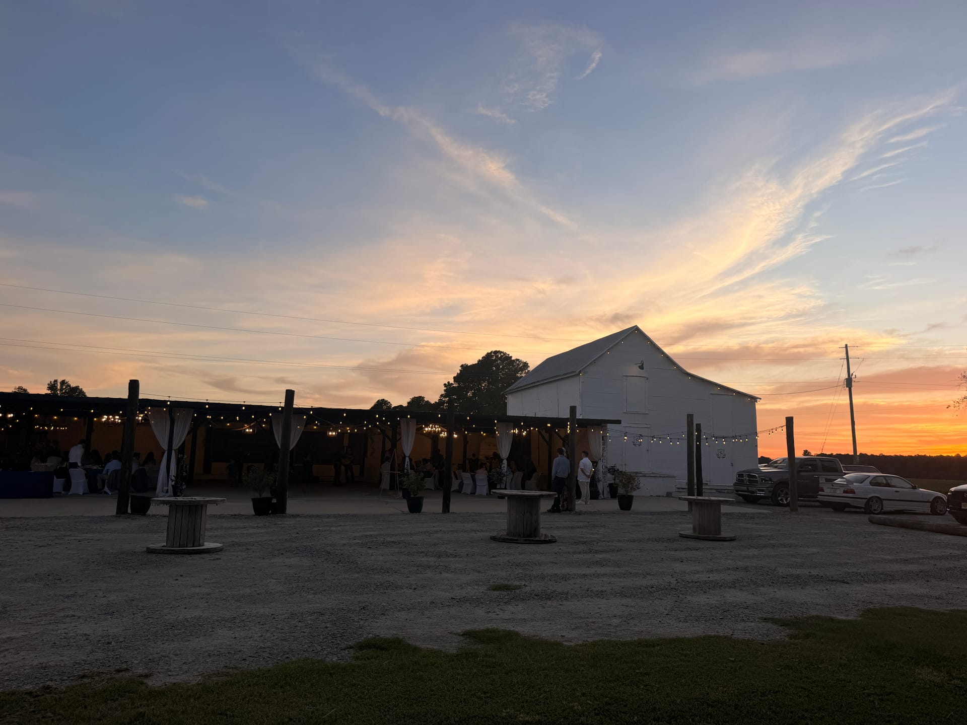 Guests mingle under string lights beside a white barn wedding venue at golden-hour sunset with warm orange and pink skies.
