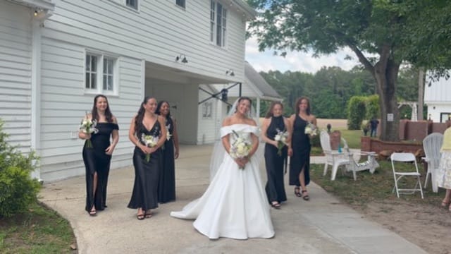 Smiling bride in a white off-shoulder ball gown stands with five bridesmaids in black dresses holding bouquets outside the historic white Scarborough House manor.