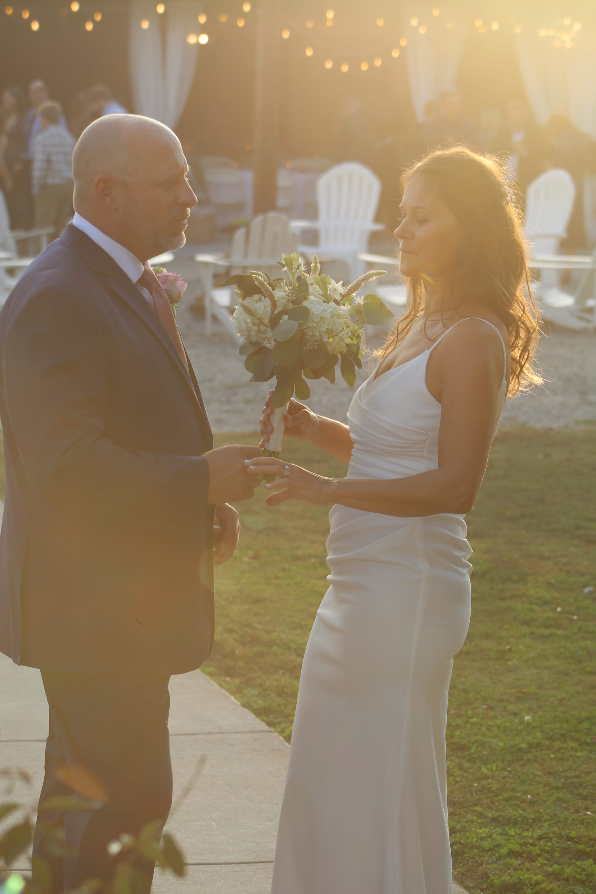 A groom places a ring on his bride's finger during an outdoor sunset ceremony on the ceremony lawn at Scarborough House, with warm golden light and string lights glowing softly in the background.