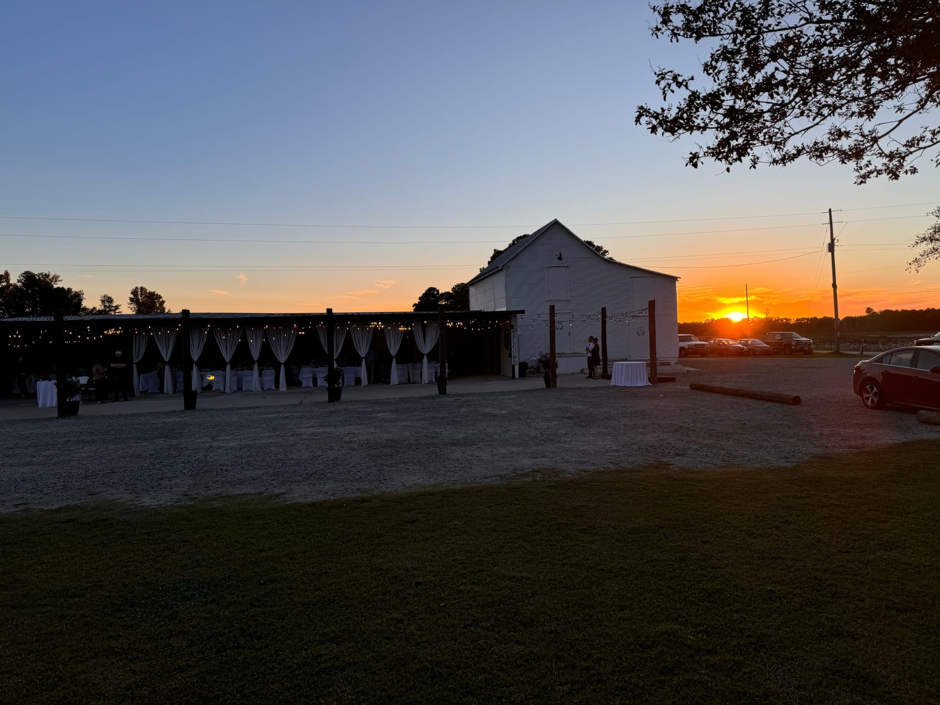 A warm golden sunset glows behind a white barn wedding venue with a draped outdoor pavilion strung with café lights at Scarborough House historic estate.