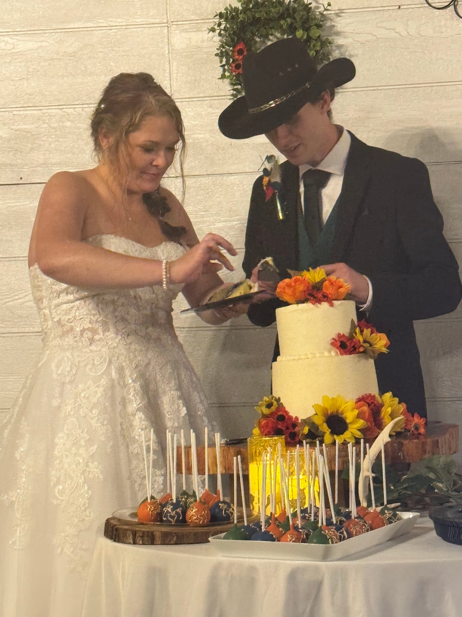 Bride in lace gown and groom in dark suit and cowboy hat cutting a two-tiered white wedding cake adorned with sunflowers and autumn blooms surrounded by decorated cake pops at Scarborough House.