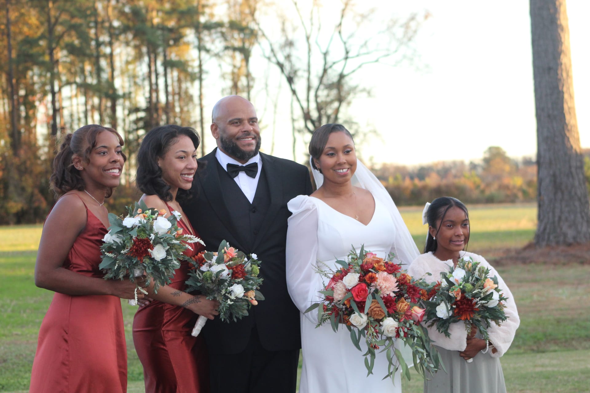 Bride in a white gown and groom in a black tuxedo pose with two bridesmaids in rust satin dresses and a young flower girl holding lush autumn bouquets on the open farmland lawn at Scarborough House.