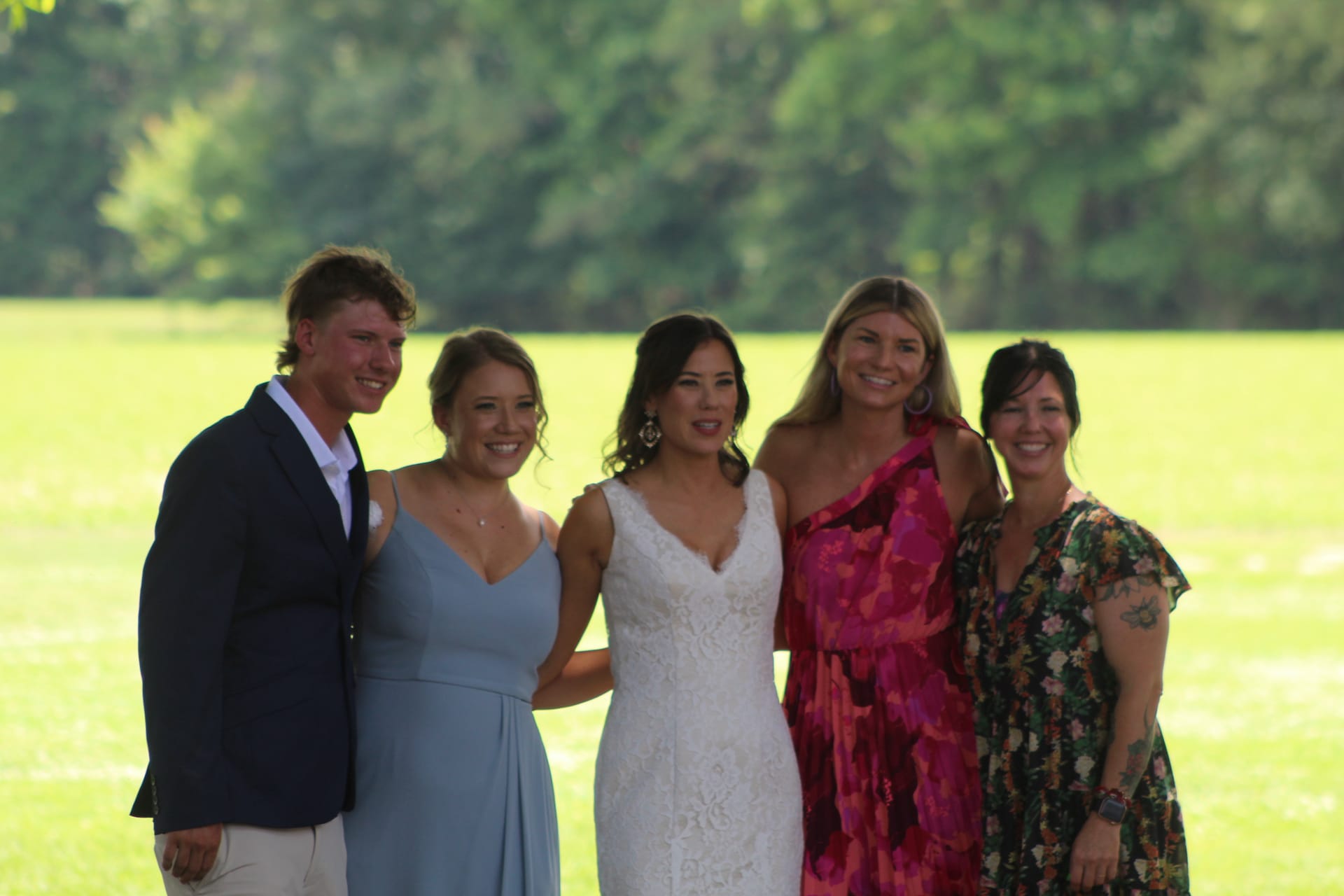 Bride in a white lace gown poses smiling with four guests dressed in colorful and blue formal attire on the sunlit lawn at Scarborough House estate.