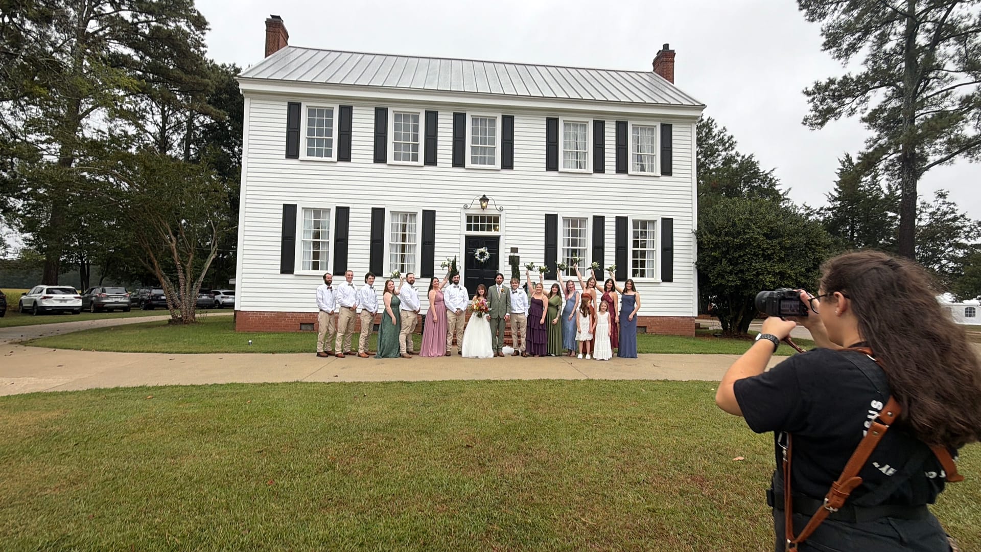 A wedding party of approximately twenty guests poses on the front steps and walkway of the white 1821 Scarborough House manor while a female photographer captures the moment from the foreground lawn.