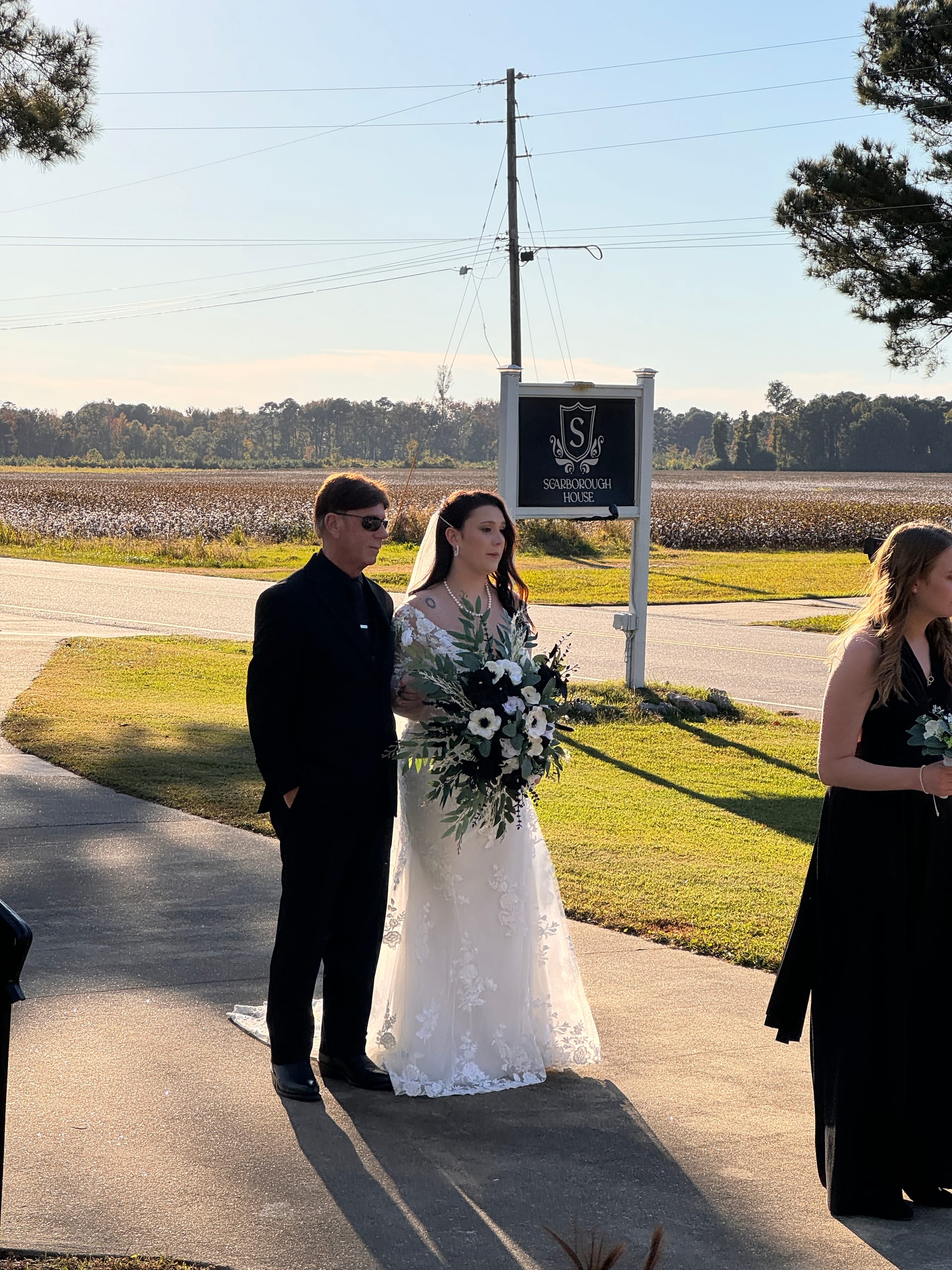 Bride in a lace gown holding a black and white floral bouquet walks with her escort past the Scarborough House marquee sign, with open cotton fields in the background on a sunny autumn day.