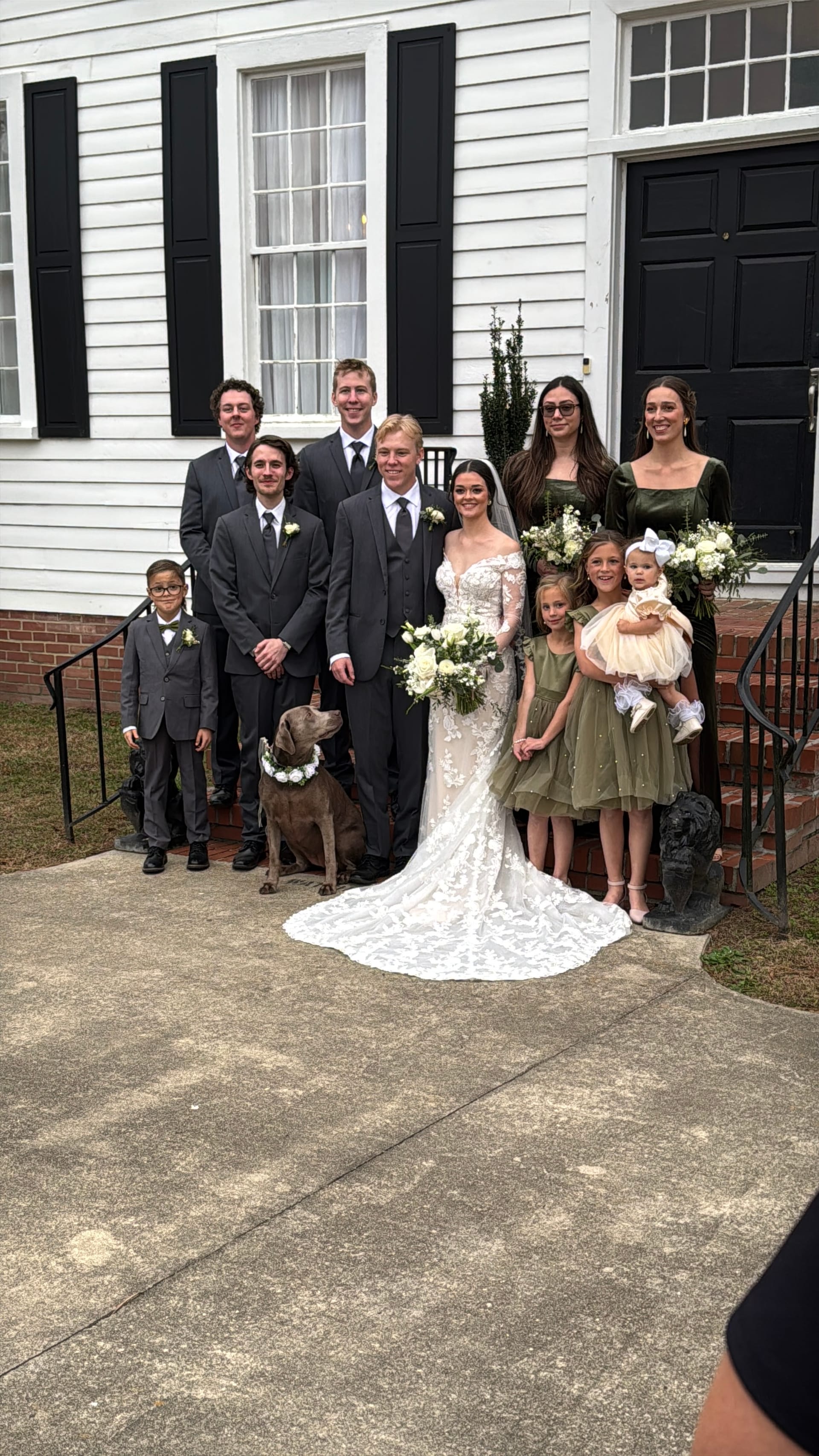 Bride and groom surrounded by their wedding party including groomsmen in charcoal suits, bridesmaids in sage green dresses, flower girls, a ring bearer, and a dog wearing a floral crown on the front steps of a white historic manor house.