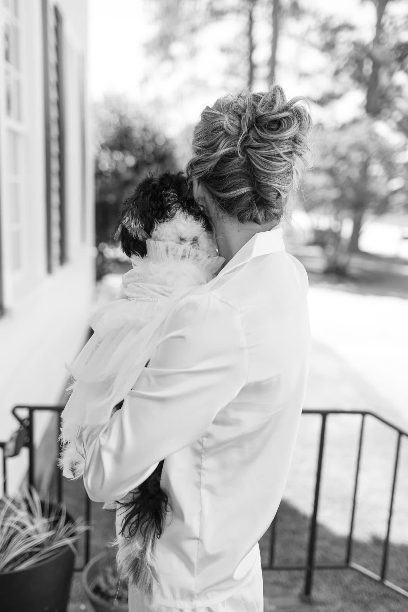 Bride in a white satin robe holds a small black and white dog dressed in a tulle outfit on the porch of the historic Scarborough House estate, captured in black and white.