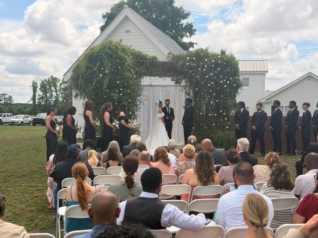 Outdoor wedding ceremony at Scarborough House with bride and groom exchanging vows beneath a lush floral arch adorned with white blooms, surrounded by bridesmaids in black gowns, groomsmen in suits, and seated guests on a sunny summer day.