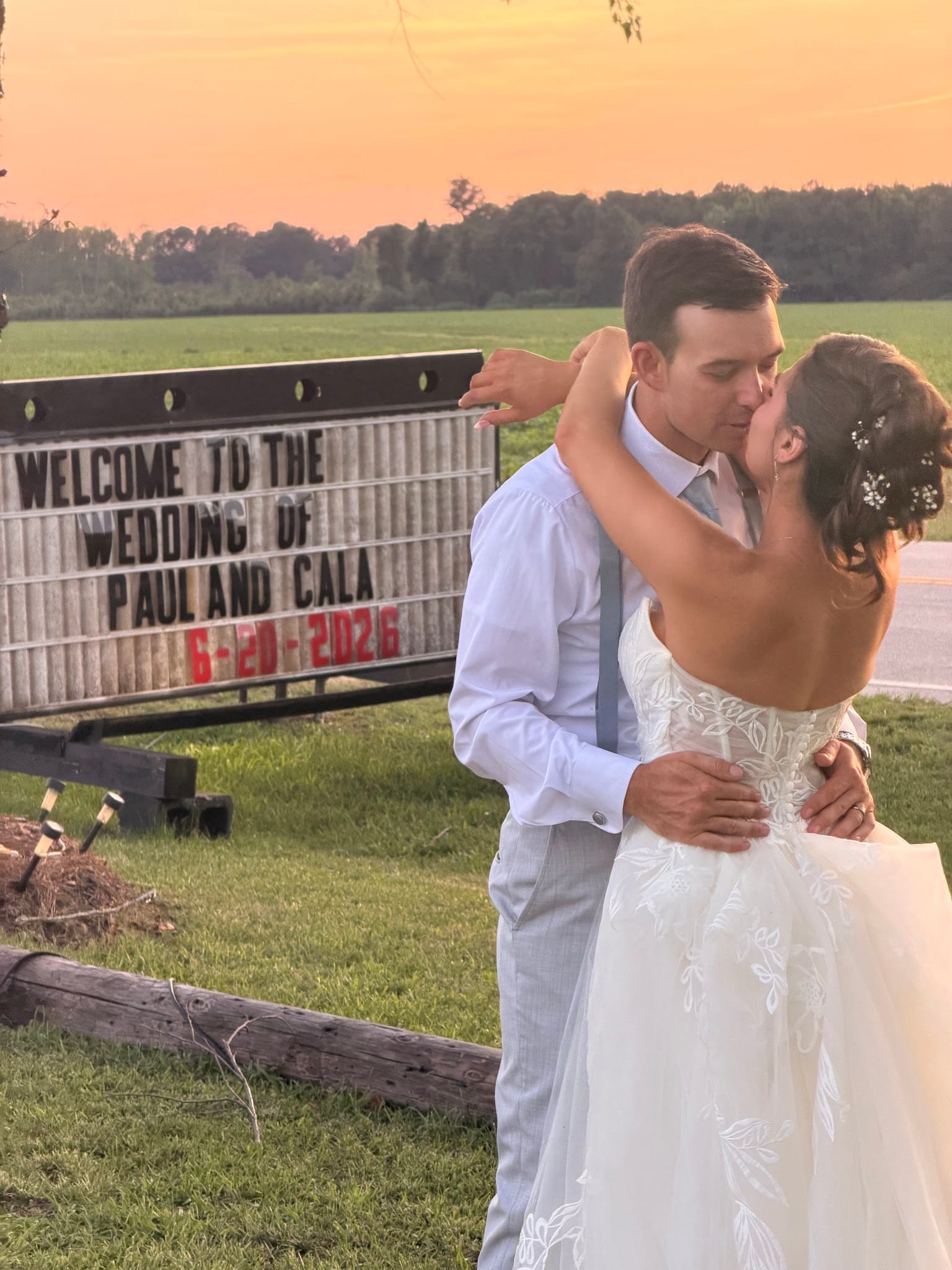Bride and groom share a kiss in front of a marquee welcome sign reading Welcome to the Wedding of Paul and Cala 6-20-2026 beneath a warm golden sunset sky at Scarborough House.