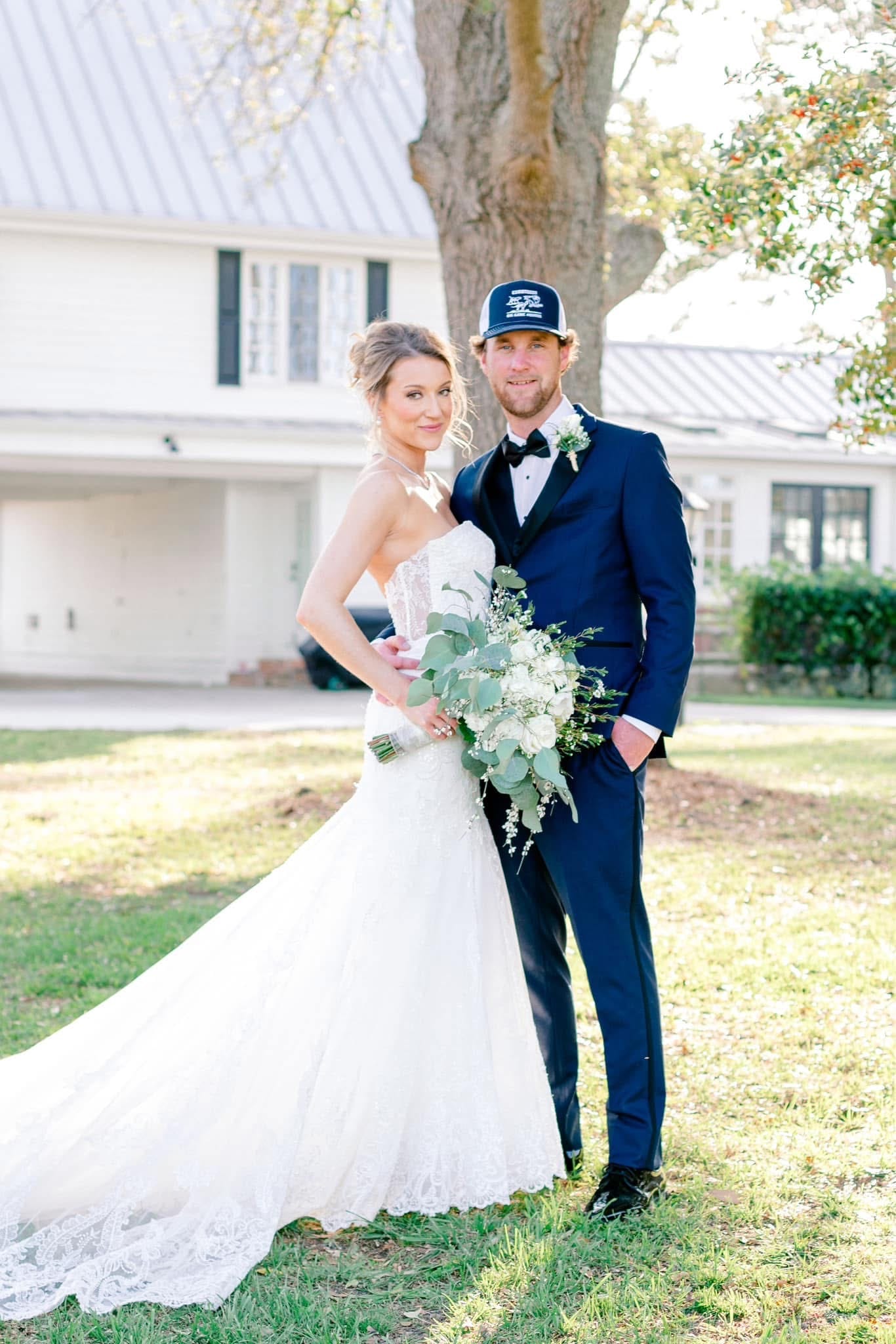 Bride in a lace strapless gown holding a lush eucalyptus and white rose bouquet stands with her groom in a navy tuxedo and trucker cap in front of the historic 1821 Scarborough House manor under a sweeping oak tree.