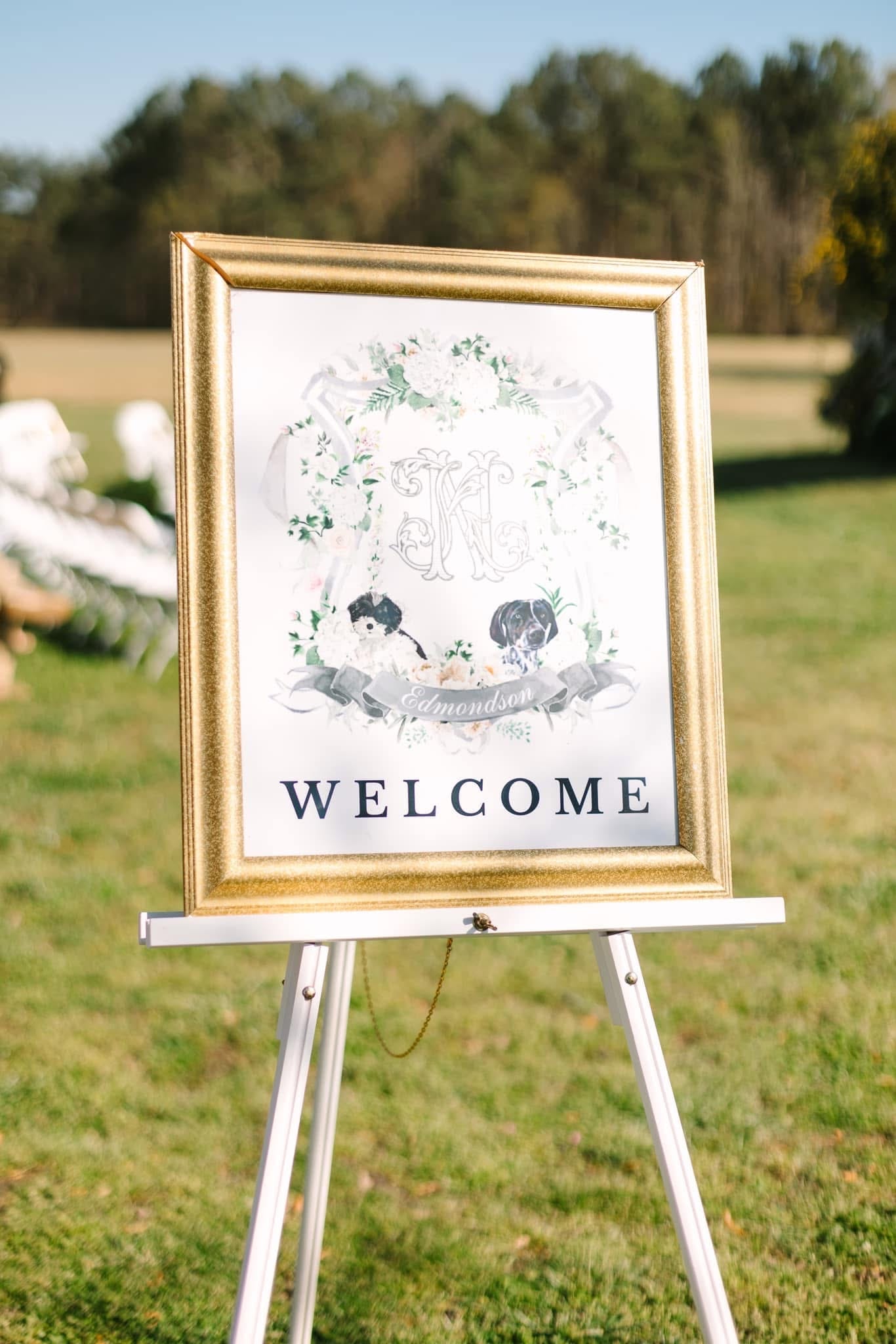A gold-framed wedding welcome sign on a white easel displaying a custom floral crest with the Edmondson monogram and illustrated dogs, set on the ceremony lawn at Scarborough House.