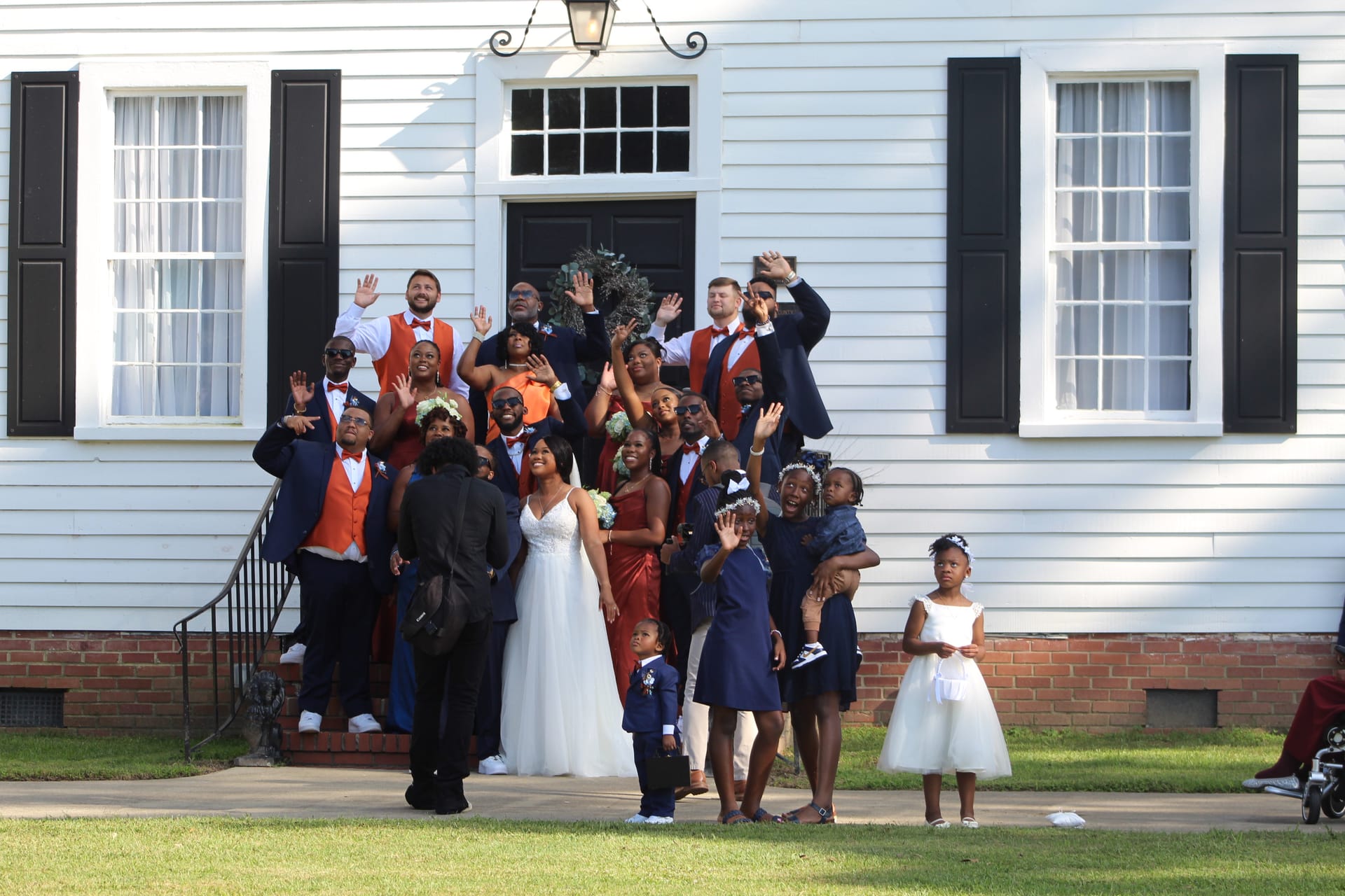 A large wedding party in navy suits and rust-orange vests waves and celebrates on the front steps of the 1821 Scarborough House manor with the bride in white at center.