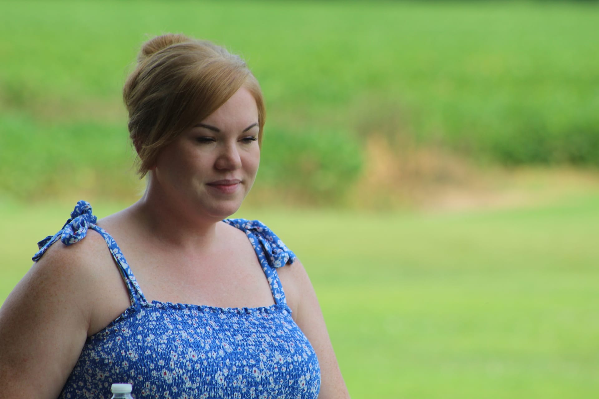 A woman with auburn hair in an updo wearing a blue floral sundress smiles softly while standing on the lush green grounds of Scarborough House estate.