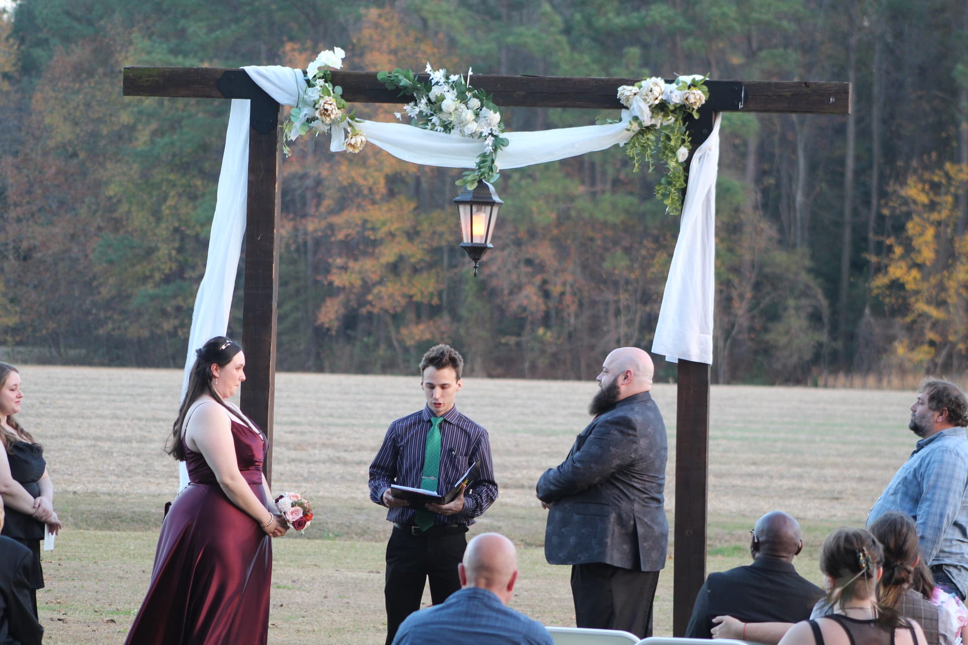 Outdoor wedding ceremony under a rustic wooden arch draped with white fabric and floral arrangements, with an officiant reading vows as guests watch across an open field framed by autumn trees.