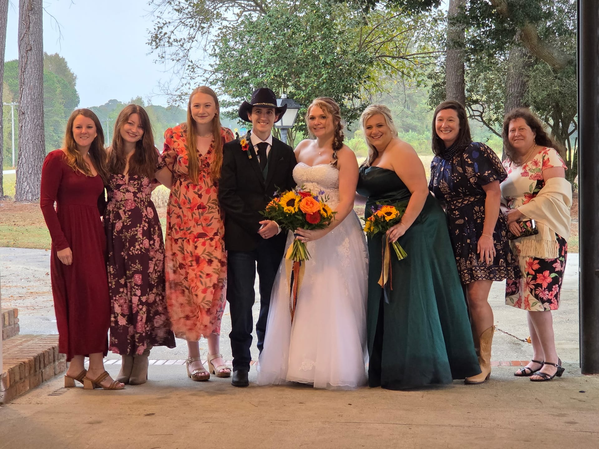Bride in a white strapless gown and groom in a black suit and cowboy hat pose with seven smiling family members holding sunflower bouquets under the pavilion at Scarborough House.