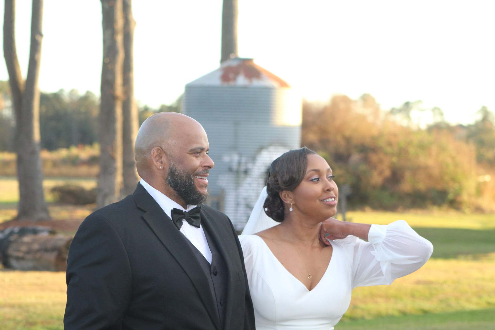 Bride in a long-sleeve white gown and groom in a black tuxedo share a relaxed candid moment on the open farmland fields of Scarborough House estate at golden hour with a grain silo and tall pines in the background.