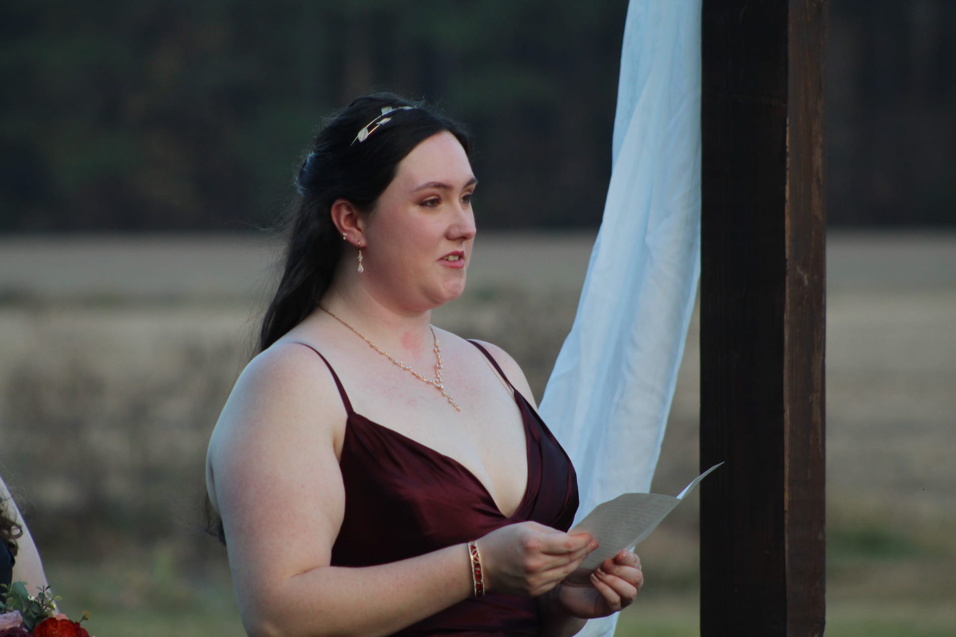 A bridesmaid in a burgundy gown reads aloud from a paper during an outdoor wedding ceremony at a historic estate venue.