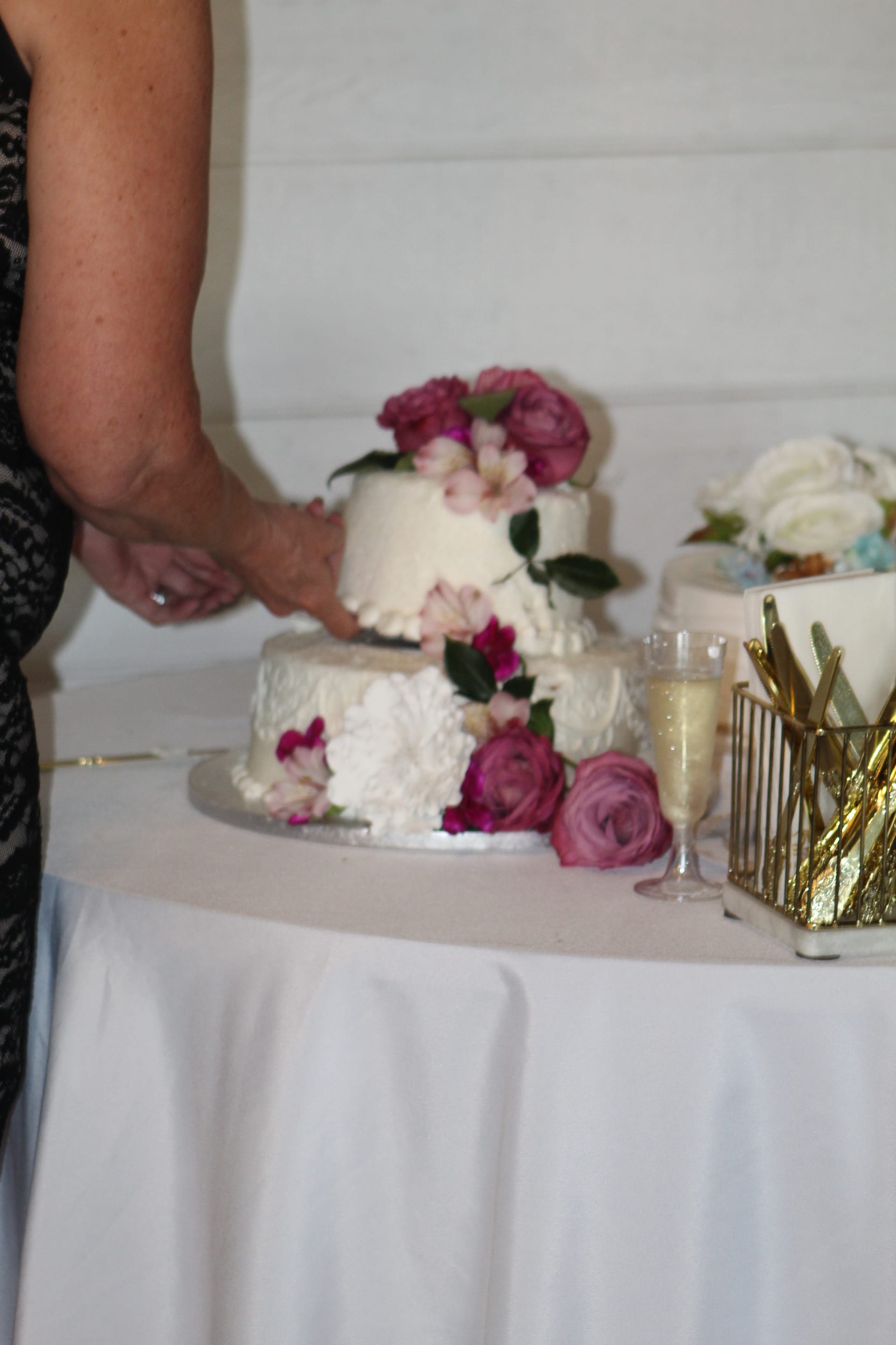 A two-tier white wedding cake decorated with fresh pink roses and peonies beside a champagne flute on a draped table at Scarborough House.