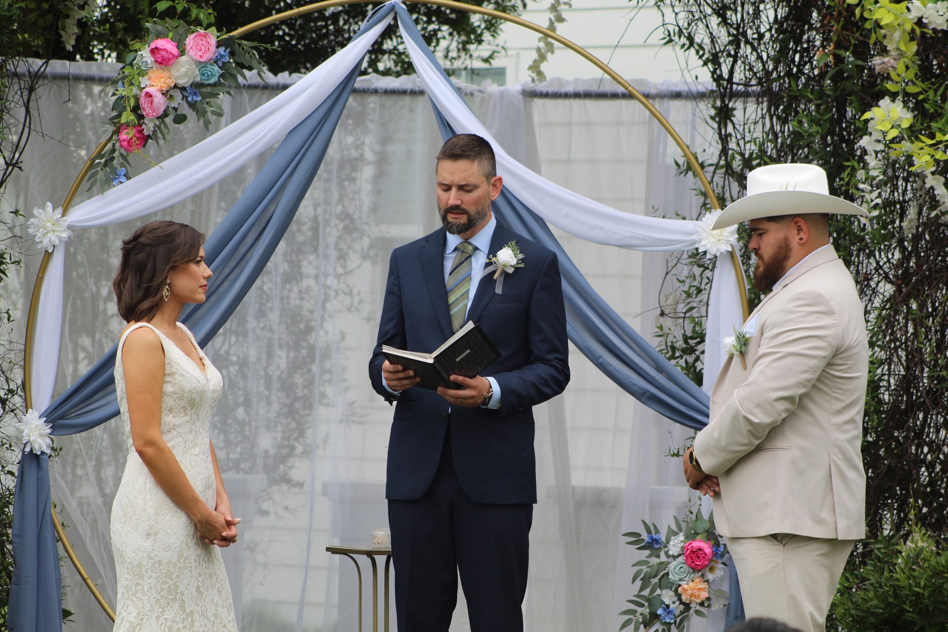Bride in lace gown and groom in tan suit with white cowboy hat exchange vows beneath a gold circular arch draped in dusty blue fabric and colorful florals at an outdoor wedding ceremony.