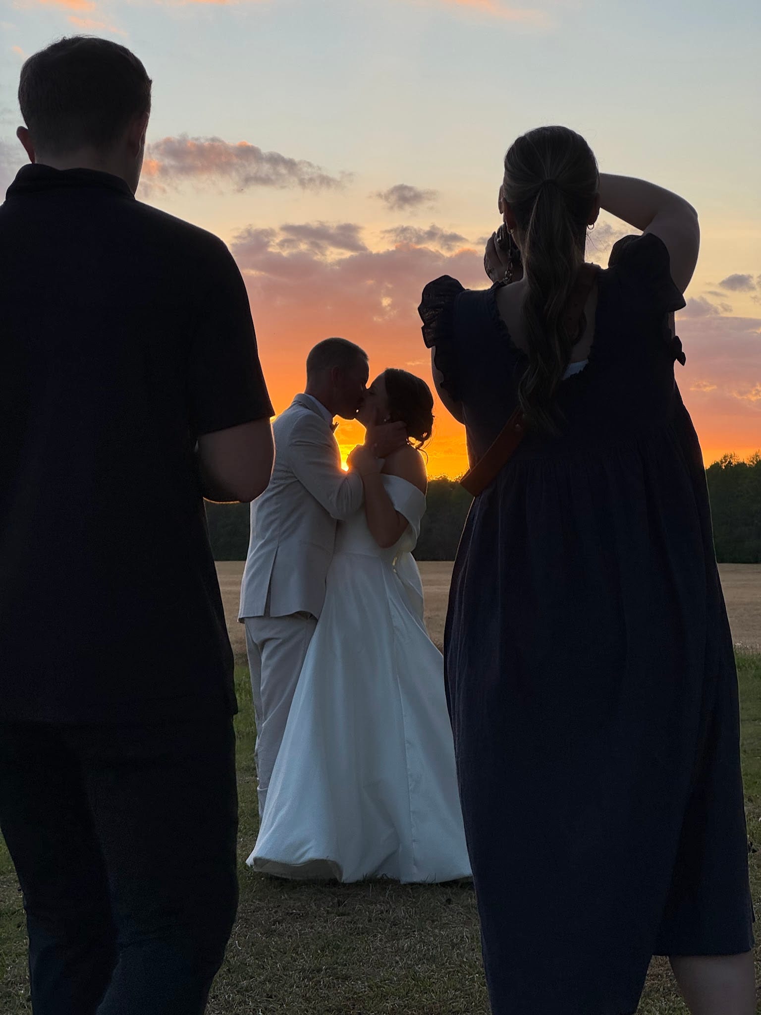 Bride and groom share a kiss silhouetted against a dramatic orange and purple sunset sky in the open farmland fields at Scarborough House, with a photographer capturing the moment in the foreground.