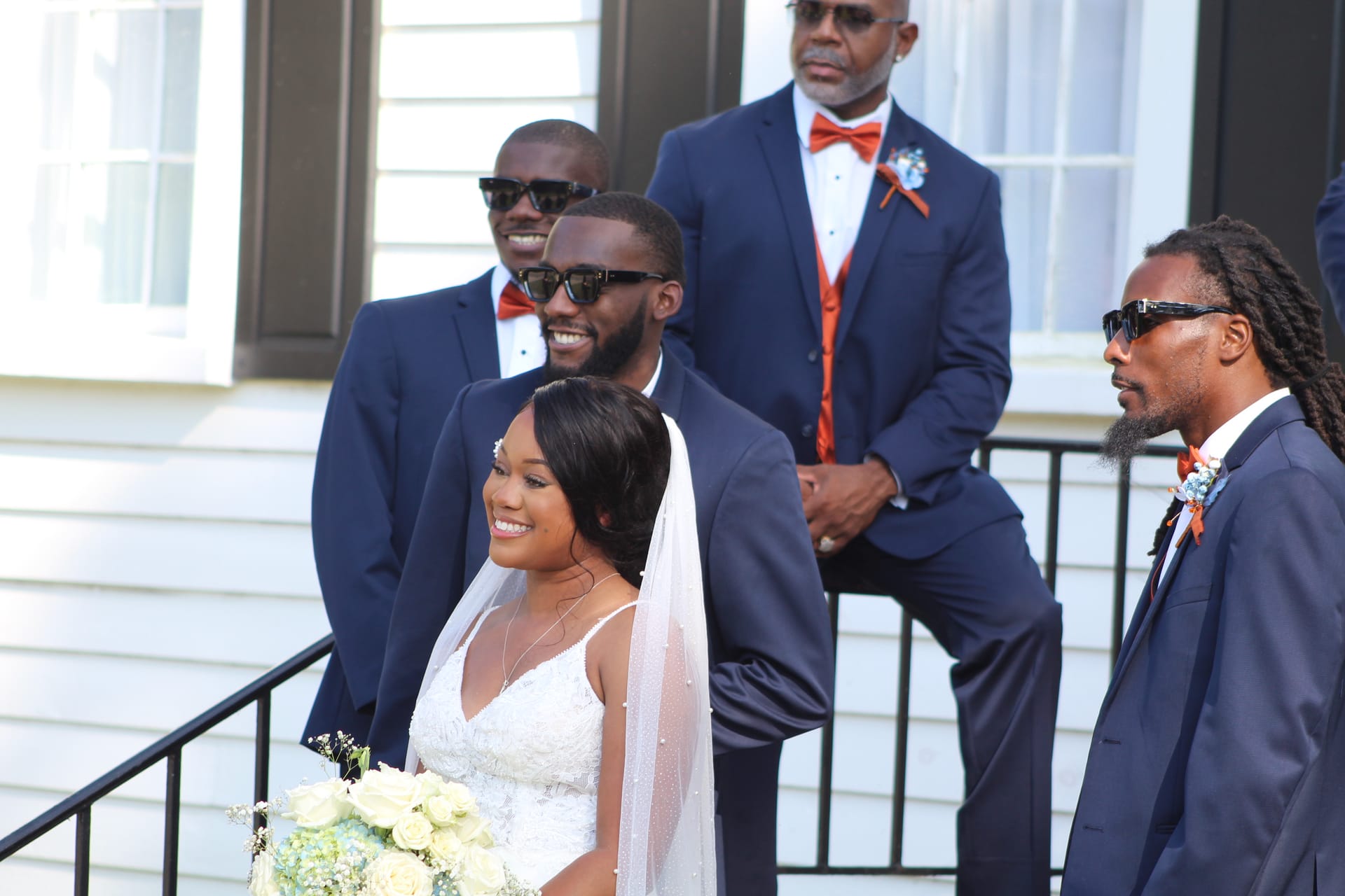 A smiling bride in a white gown holding a white floral bouquet stands on the front steps of the historic Scarborough House manor surrounded by groomsmen in navy suits with burnt orange bow ties.