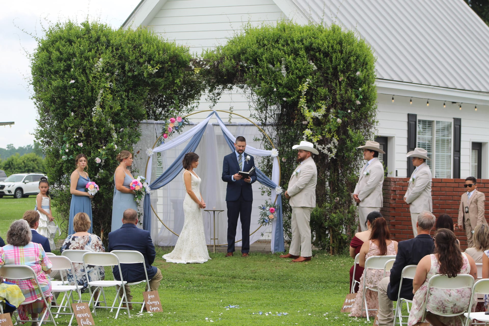 Bride and groom exchange vows beneath a gold hoop arch draped in blue fabric and flowers on the ceremony lawn at Scarborough House, with bridesmaids in blue and groomsmen in tan suits flanking the couple before a white historic chapel.