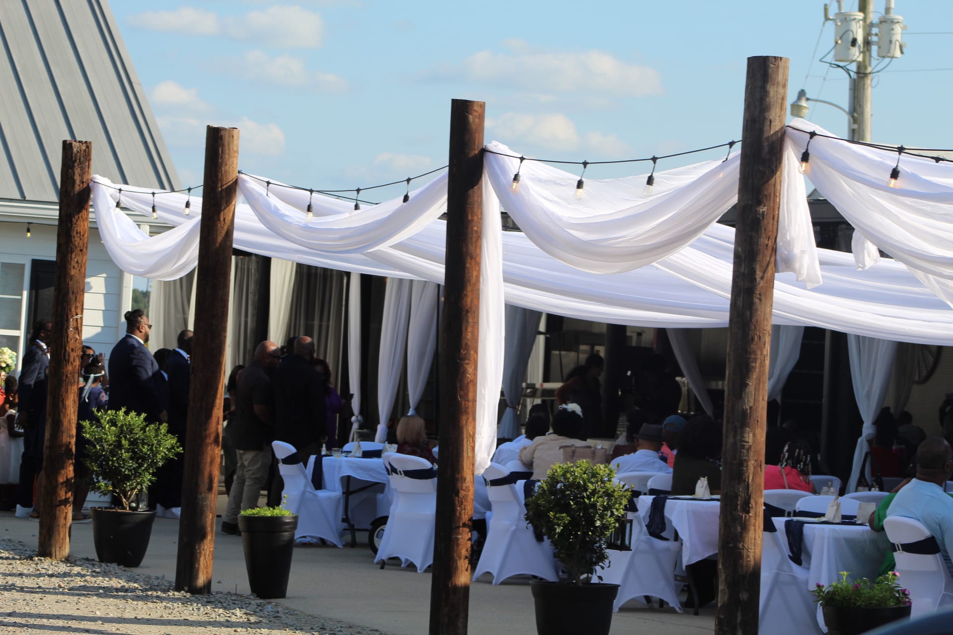 Elegant outdoor wedding reception at Scarborough House featuring white fabric draping strung between rustic wooden posts with bistro string lights, white chair-covered seating, and topiary centerpieces under a partly cloudy sky.