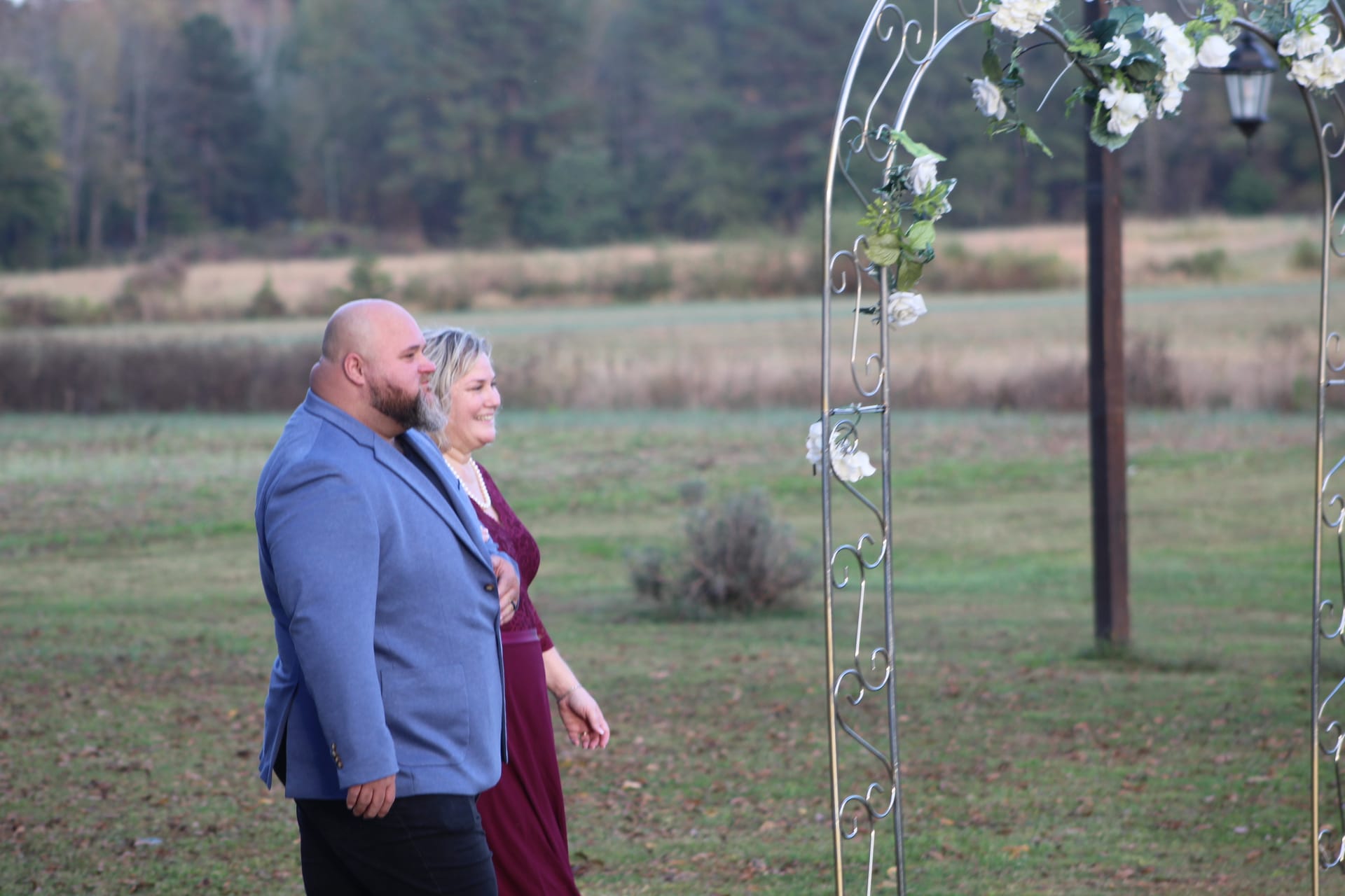 A smiling couple stands arm in arm beside a white-flower-adorned metal arch on the open ceremony lawn at Scarborough House, with rolling farmland fields and tree line visible in the background.