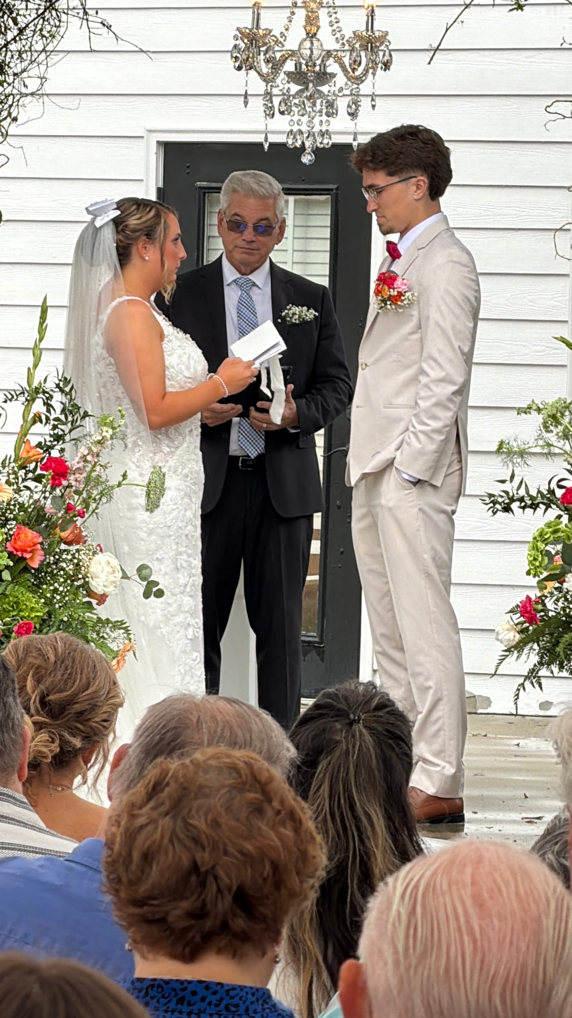 Bride and groom exchange vows before an officiant on the front steps of the historic Scarborough House estate, framed by lush colorful floral arrangements and a crystal chandelier.