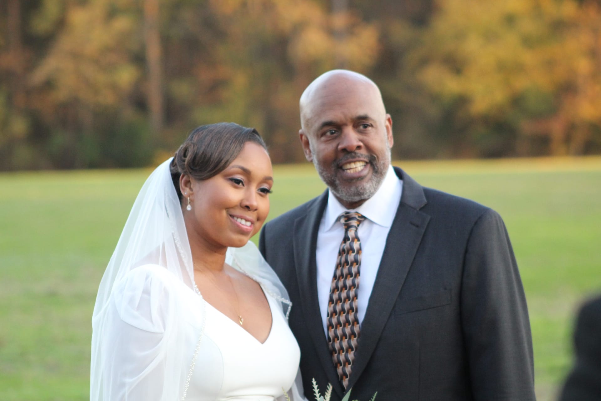 A smiling bride in a white gown and veil poses with a gentleman in a dark suit and patterned tie on the ceremony lawn surrounded by golden autumn trees at Scarborough House.