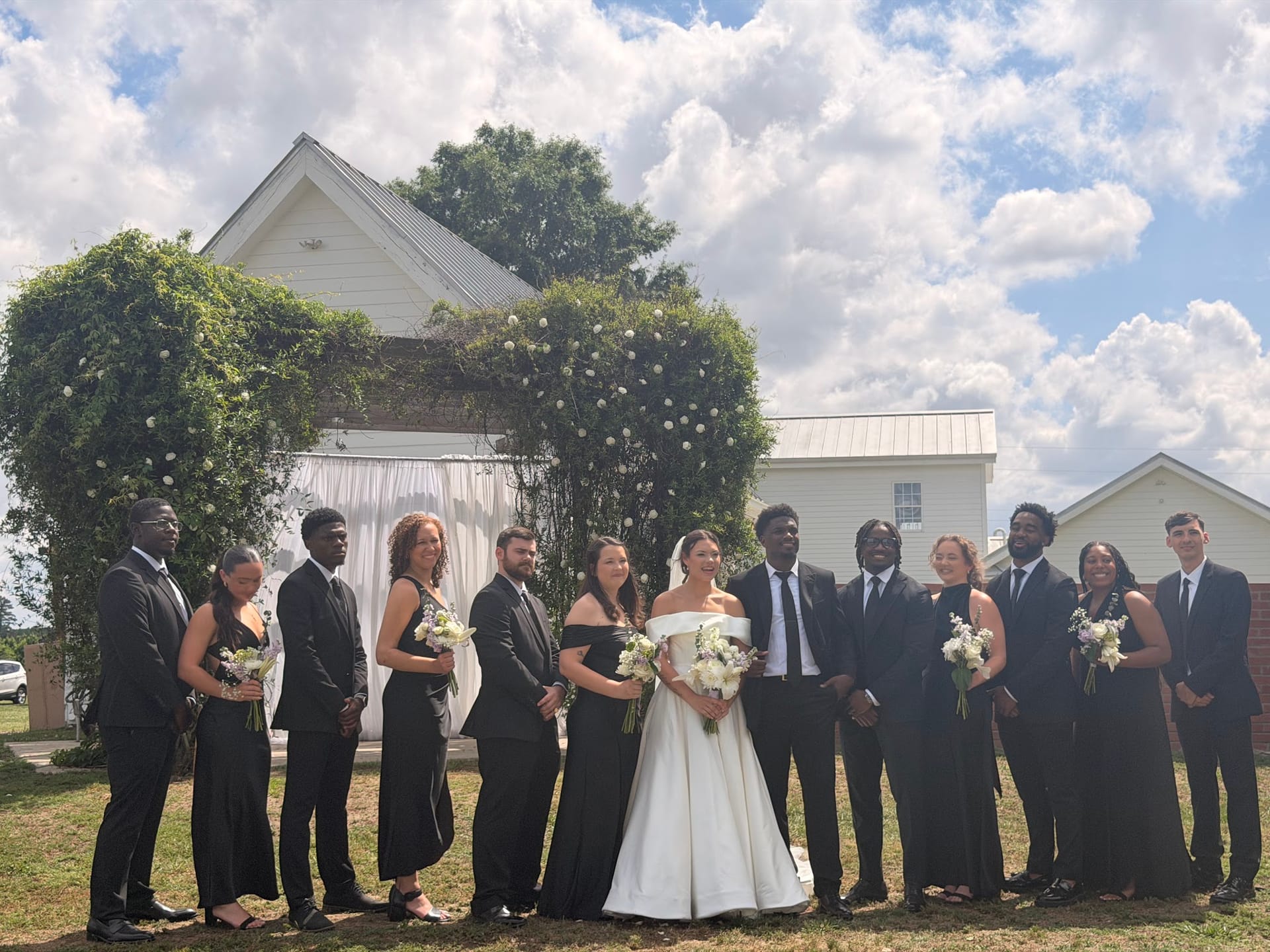 Bride and groom surrounded by their wedding party in coordinating black attire posing in front of an ivy-covered white chapel on the ceremony lawn at Scarborough House.