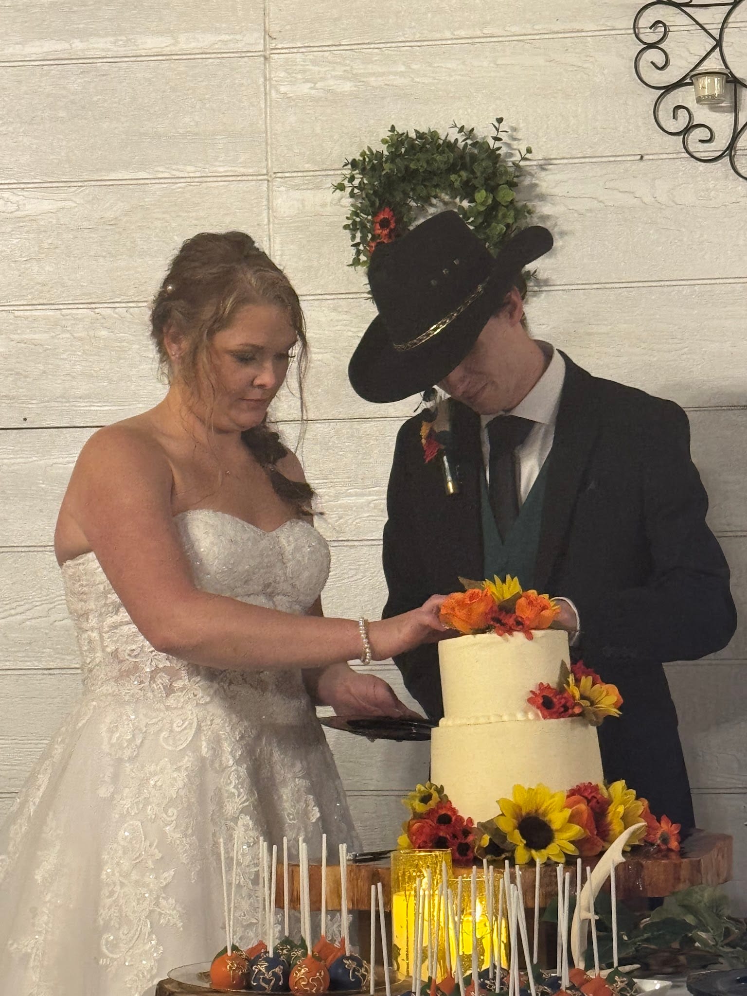 Bride in lace gown and groom in black suit and cowboy hat cut their two-tier white wedding cake adorned with sunflowers and autumn blooms at a fall wedding reception.