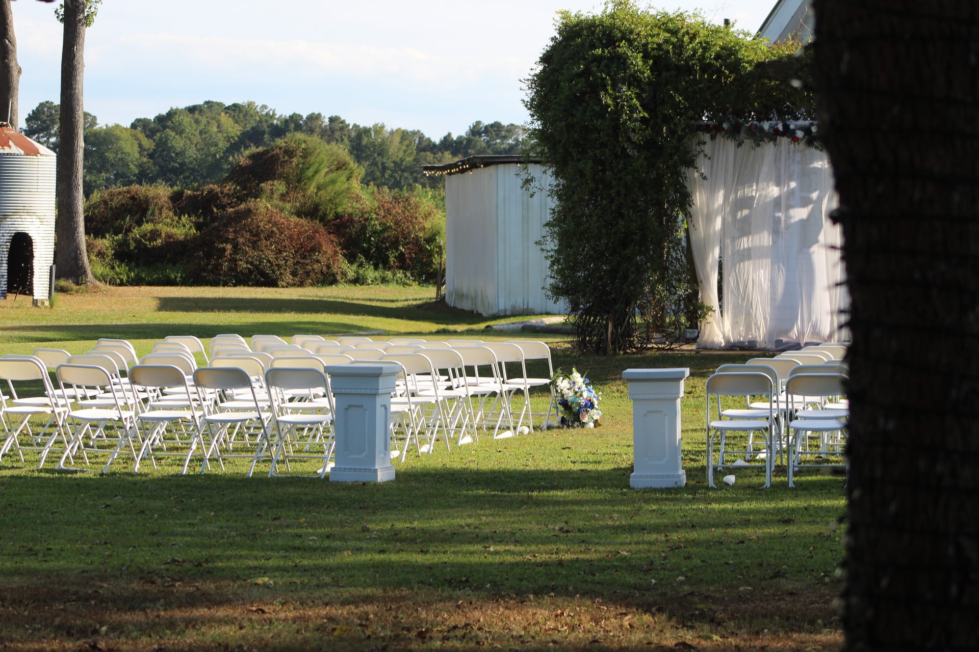 Rows of white folding chairs arranged along a grassy ceremony lawn with white pedestal columns, a floral arrangement, and a draped backdrop at Scarborough House outdoor wedding venue.