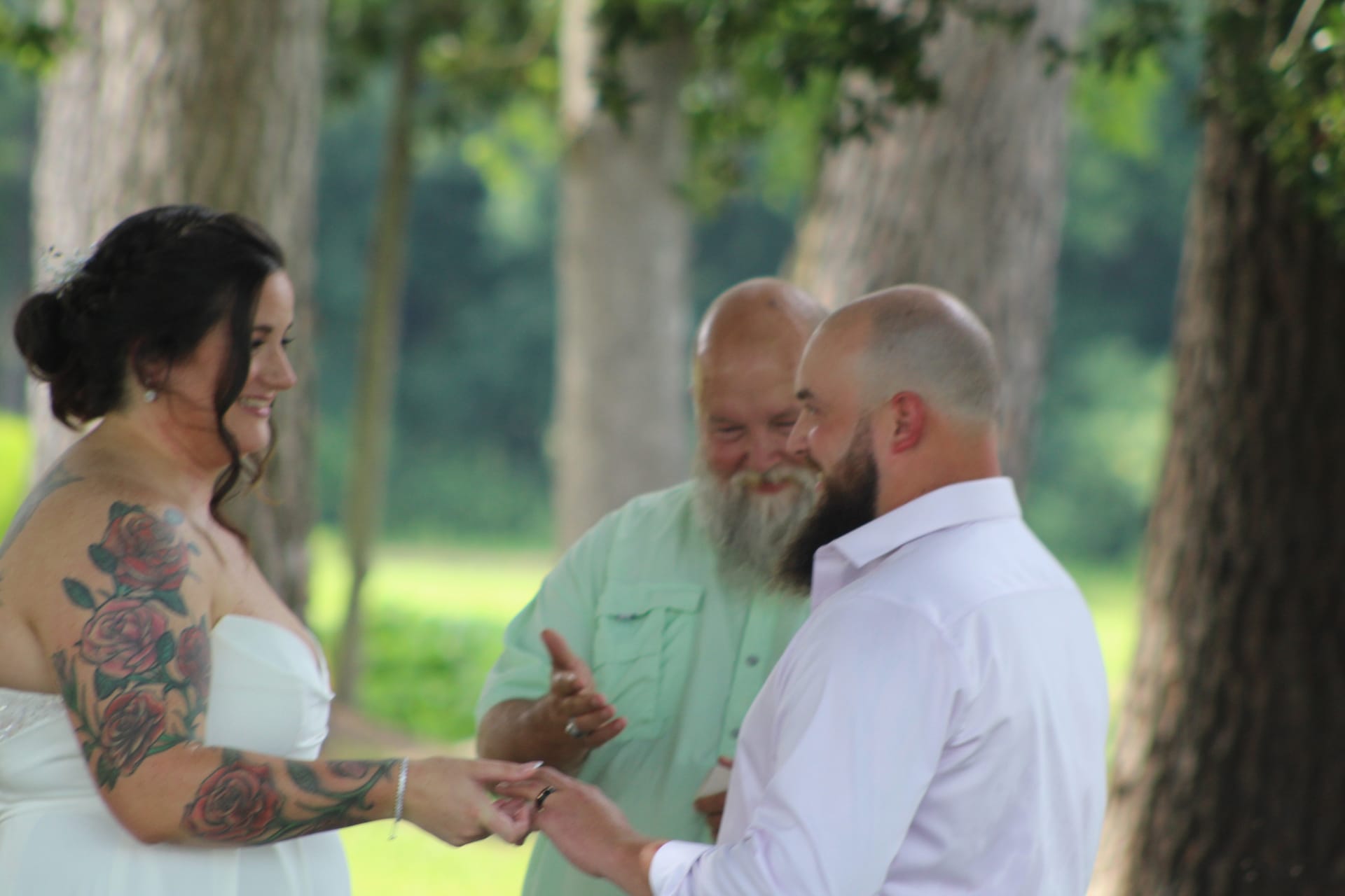 Tattooed bride in white strapless gown smiles as groom places ring on her finger during an outdoor wedding ceremony beneath tall oak trees at a historic estate venue.