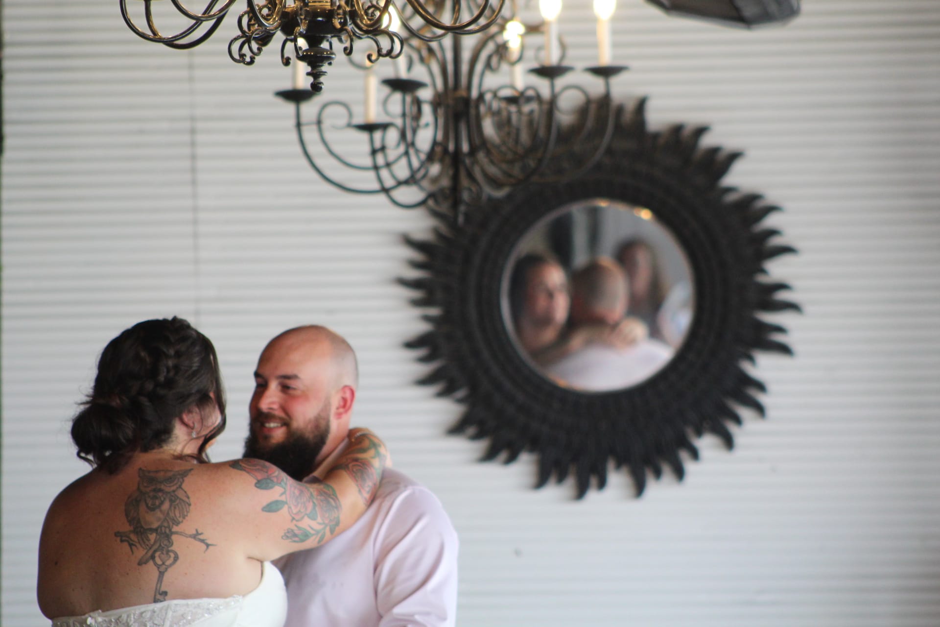A tattooed bride and smiling bearded groom share an intimate dance in Scarborough Houses grand hall beneath an ornate chandelier with their reflection captured in a decorative sunburst mirror on the white shiplap wall.