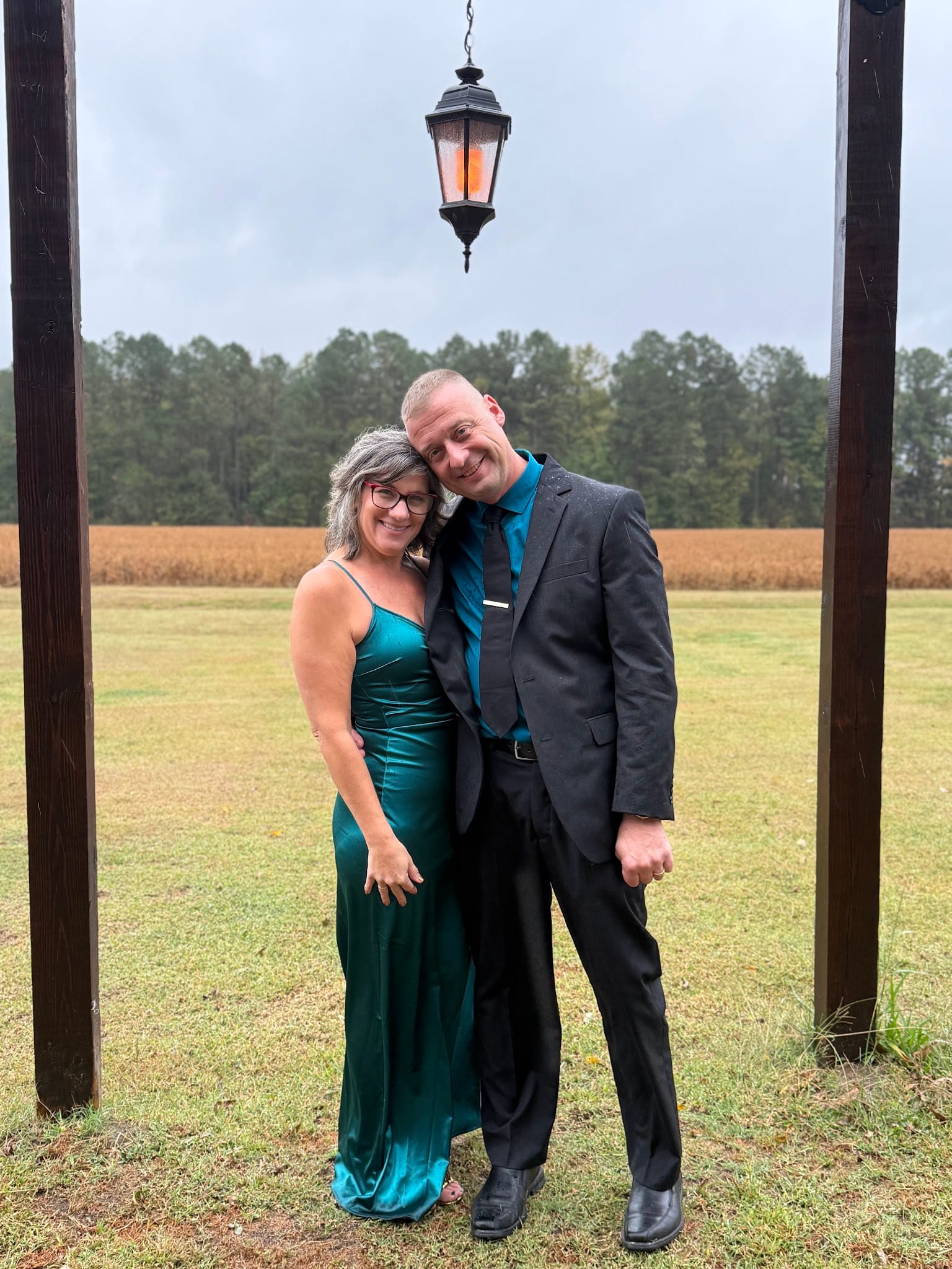 A smiling couple dressed in a teal velvet gown and charcoal suit pose together beneath a wooden arch with a glowing lantern, surrounded by open farmland fields at Scarborough House.