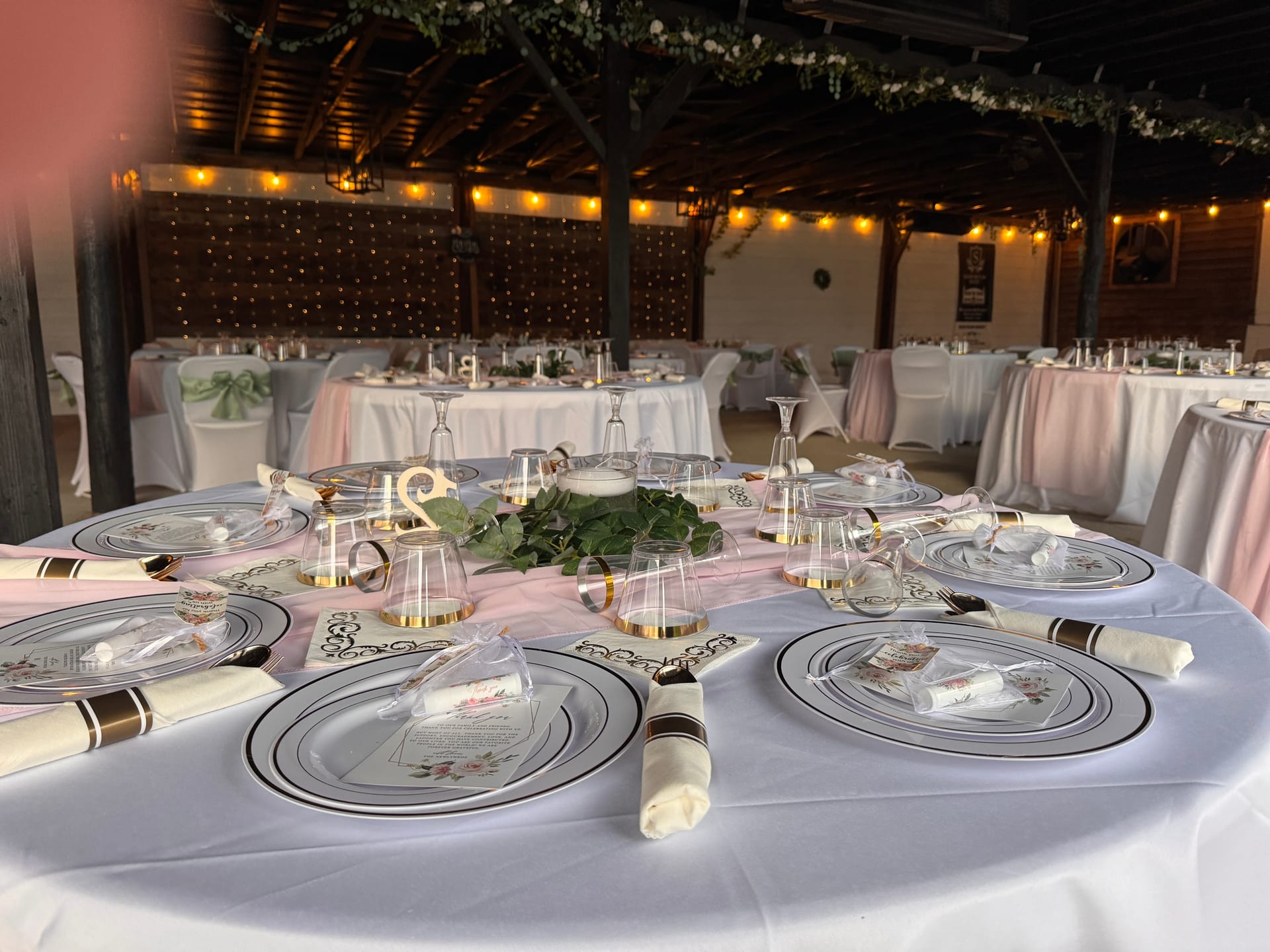 Elegantly set round reception table with white linen, gold-rimmed charger plates, clear glassware, and a lush greenery centerpiece under warm string lights at Scarborough House magnolia pavilion.