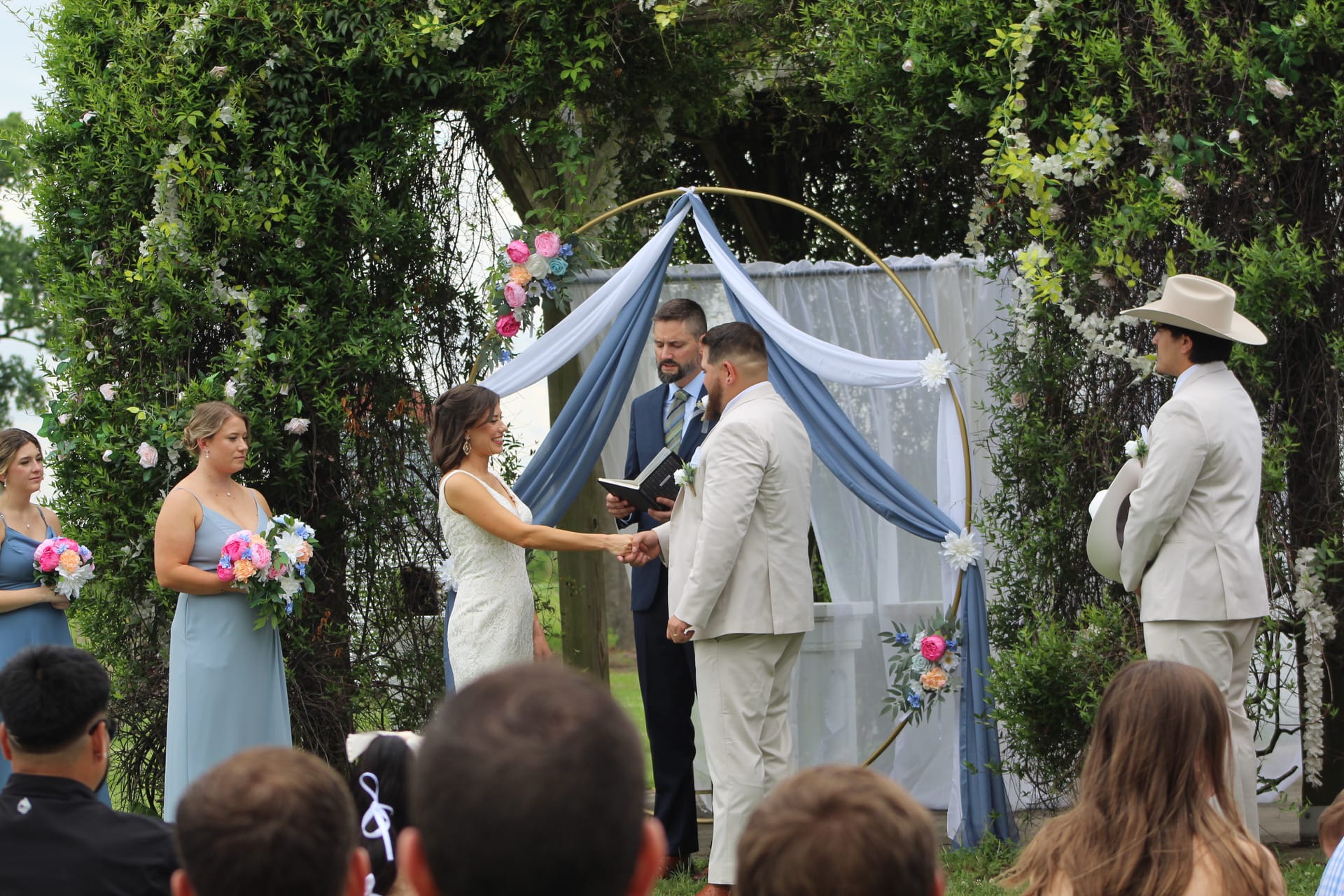 Bride and groom hold hands while exchanging vows beneath a gold circular arch draped in blue fabric and colorful flowers, flanked by bridesmaids and groomsmen on a lush outdoor ceremony lawn.
