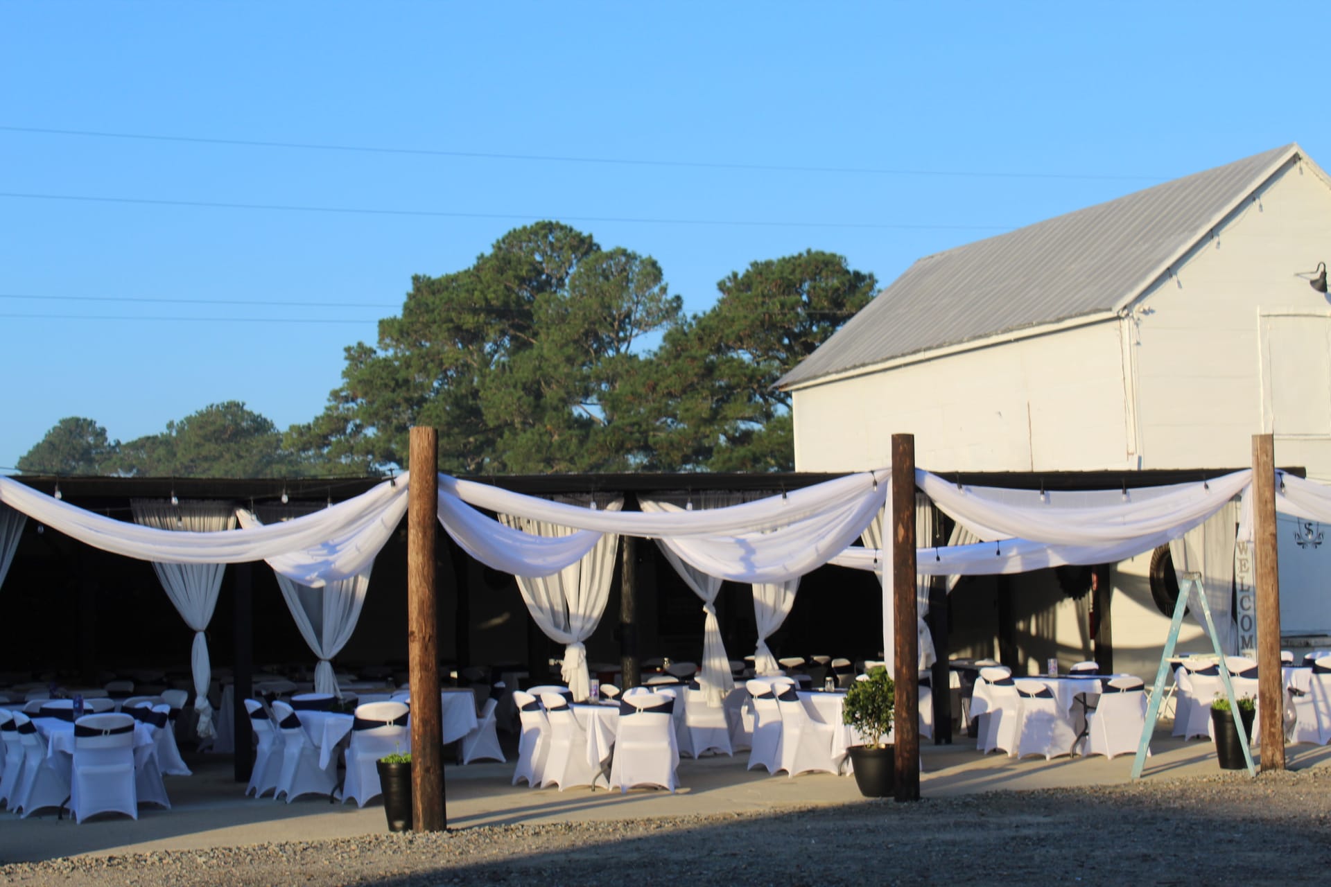 Outdoor wedding reception area at Scarborough House featuring white draped fabric swags over rustic wooden posts with white linen-covered tables and chairs set for guests in warm evening light.