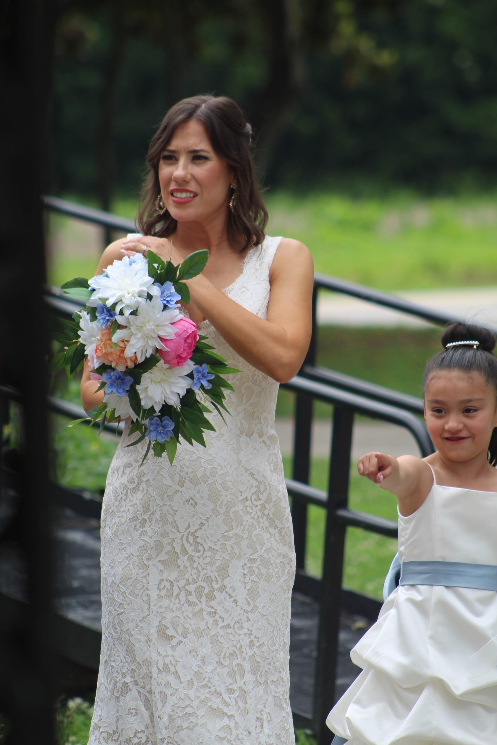 A smiling bride in a lace wedding gown holds a pink white and blue bouquet while walking alongside a young flower girl in a white dress with a blue sash at an outdoor wedding venue.