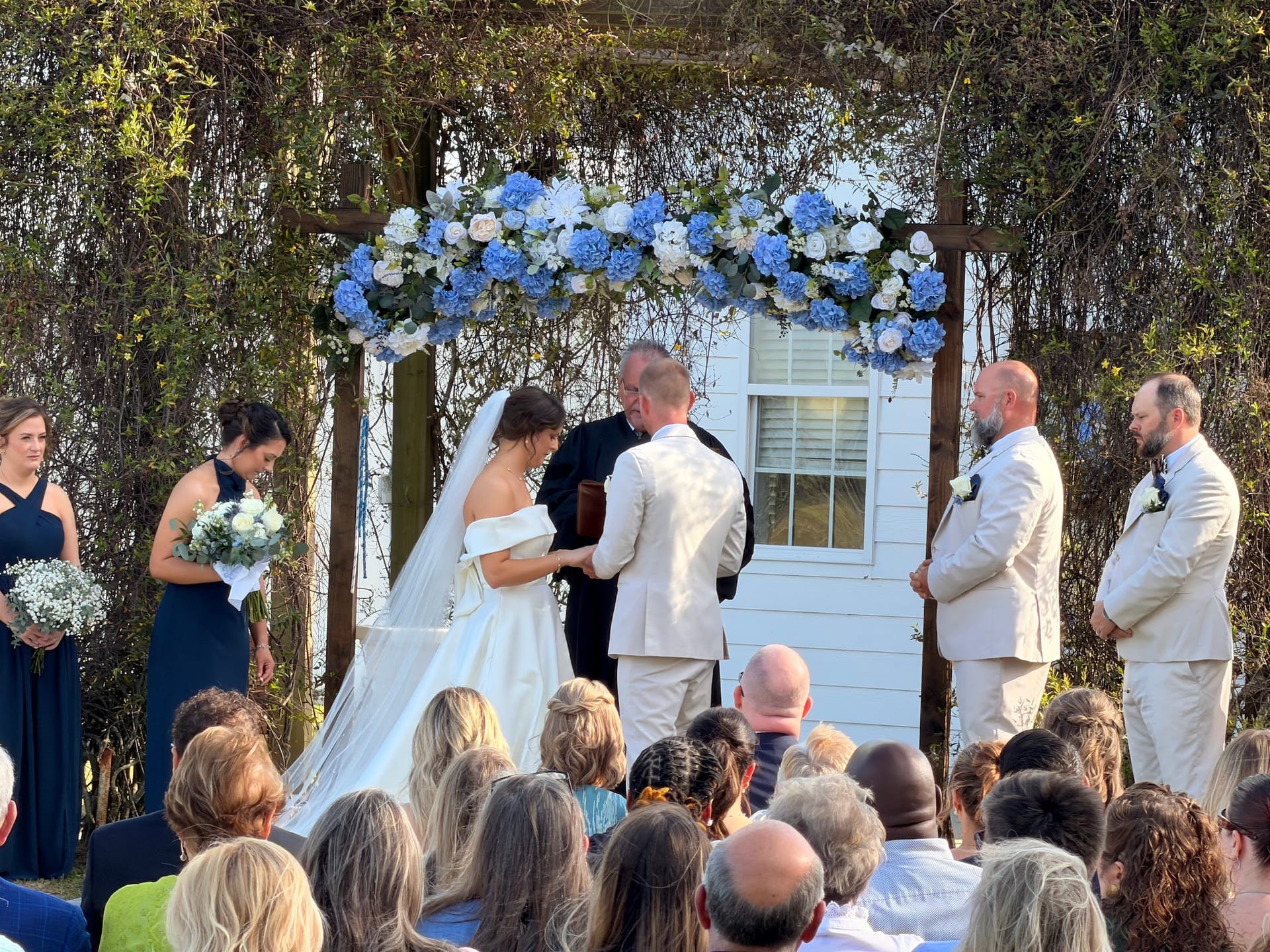 Bride and groom exchange vows beneath a wooden arbor adorned with blue hydrangeas and white roses on the ceremony lawn at Scarborough House, surrounded by bridesmaids in navy, groomsmen in ivory, and seated guests.