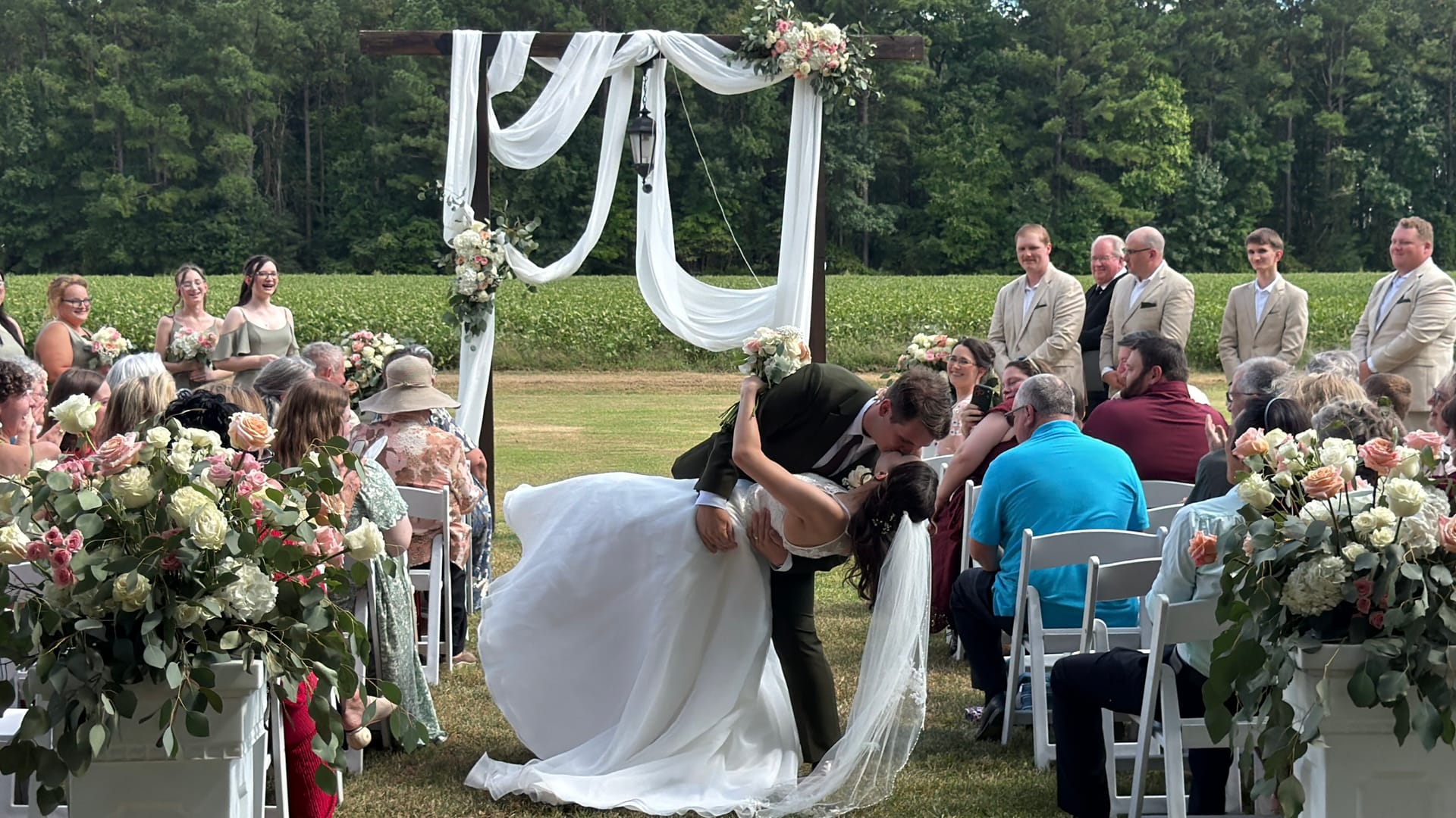 Groom dips and kisses his bride beneath a white-draped wooden arch adorned with blush florals on the ceremony lawn at Scarborough House as delighted guests look on.